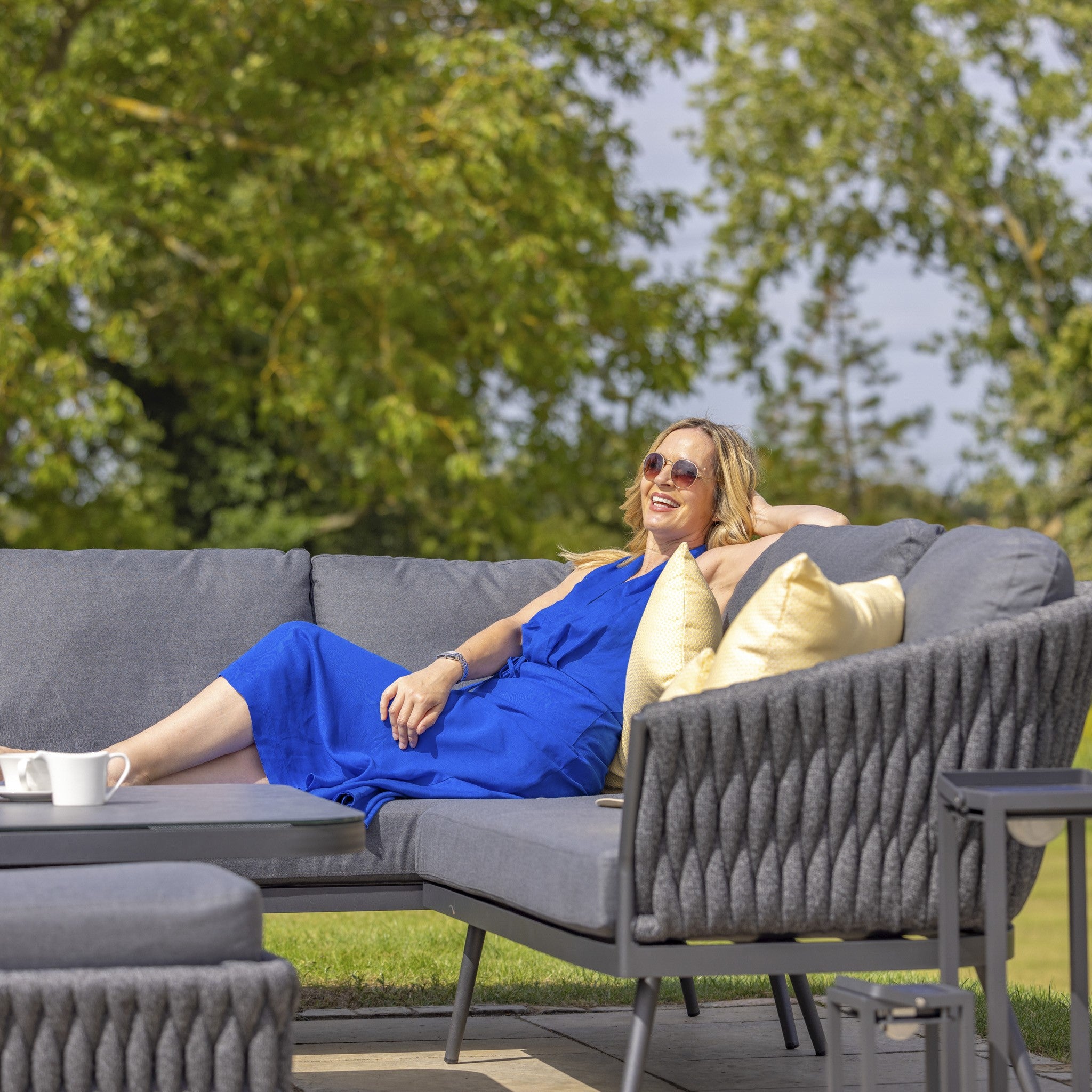 A woman in a blue dress lounges on an outdoor sofa, part of the stylish Palma Rope Rectangular Corner Dining Set with Rising Table in Grey (Right Hand), featuring a water-repelling thick rope weave and durable aluminium frames, smiling and enjoying the sunny day with trees in the background.