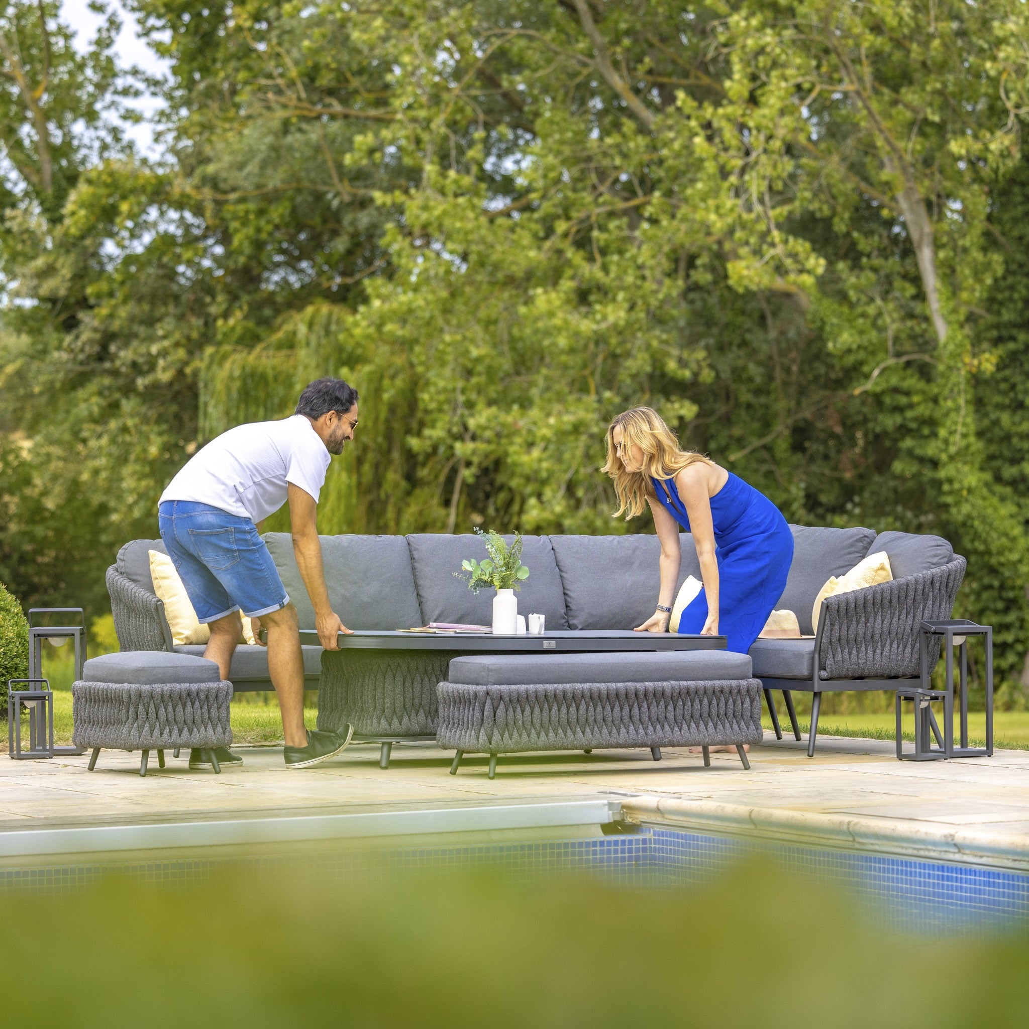 Two people rearrange the Palma Rope Rectangular Corner Dining Set with Rising Table in Grey (Right Hand) on a patio near a pool, with green trees in the background. The water-repelling thick rope weave and sturdy aluminium frames ensure that this outdoor furniture can withstand any weather while adding style to their relaxing space.