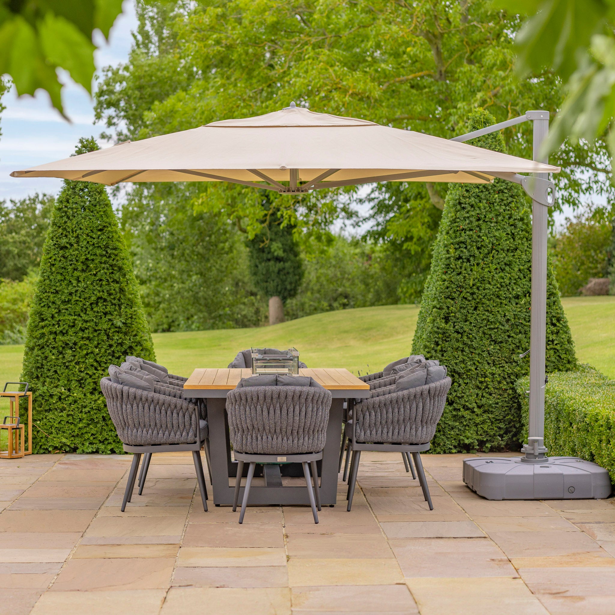 Outdoor dining area with a wooden table, gray chairs, and a large Pallas 4m x 3m Rectangular Cantilever Parasol with LED Lighting in Beige, surrounded by greenery and trimmed bushes.