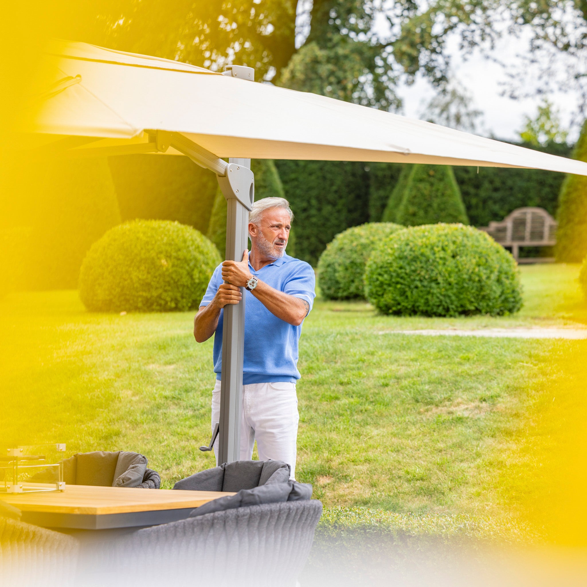 A man in a blue shirt and white pants adjusting the Pallas 4m x 3m Rectangular Cantilever Parasol with LED Lighting in Beige in a garden with neatly trimmed bushes, providing perfect garden shade.