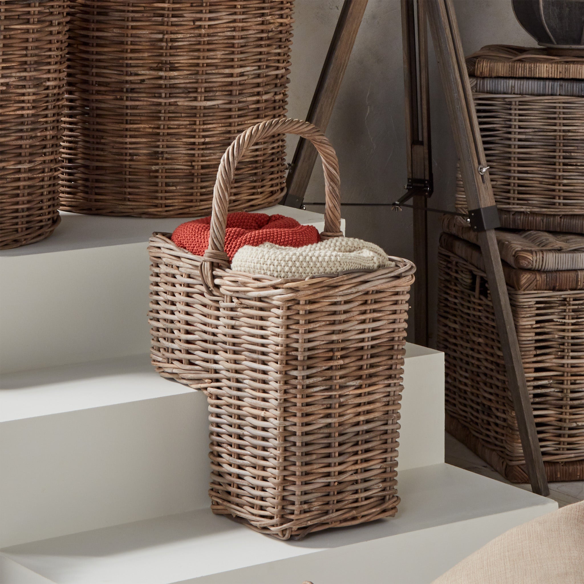 The Bali Grey Kubu Stair Basket by Pacific Lifestyle displays folded red and beige knitted blankets on a white stepped surface.