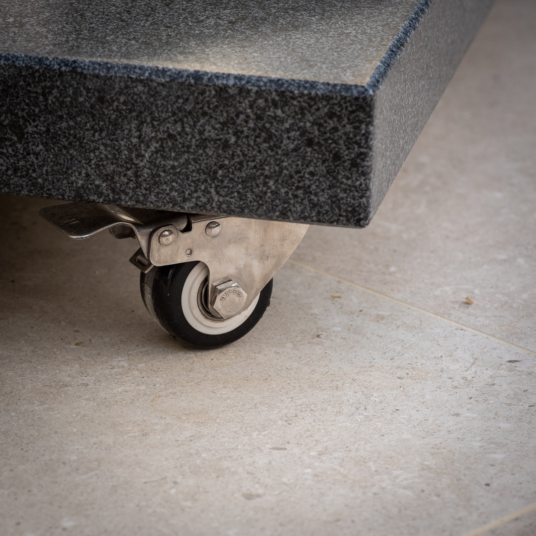 Close-up of a metal swivel caster wheel attached to the Granite 90kg Cantilever Parasol Base With Wheels on a beige tiled floor.