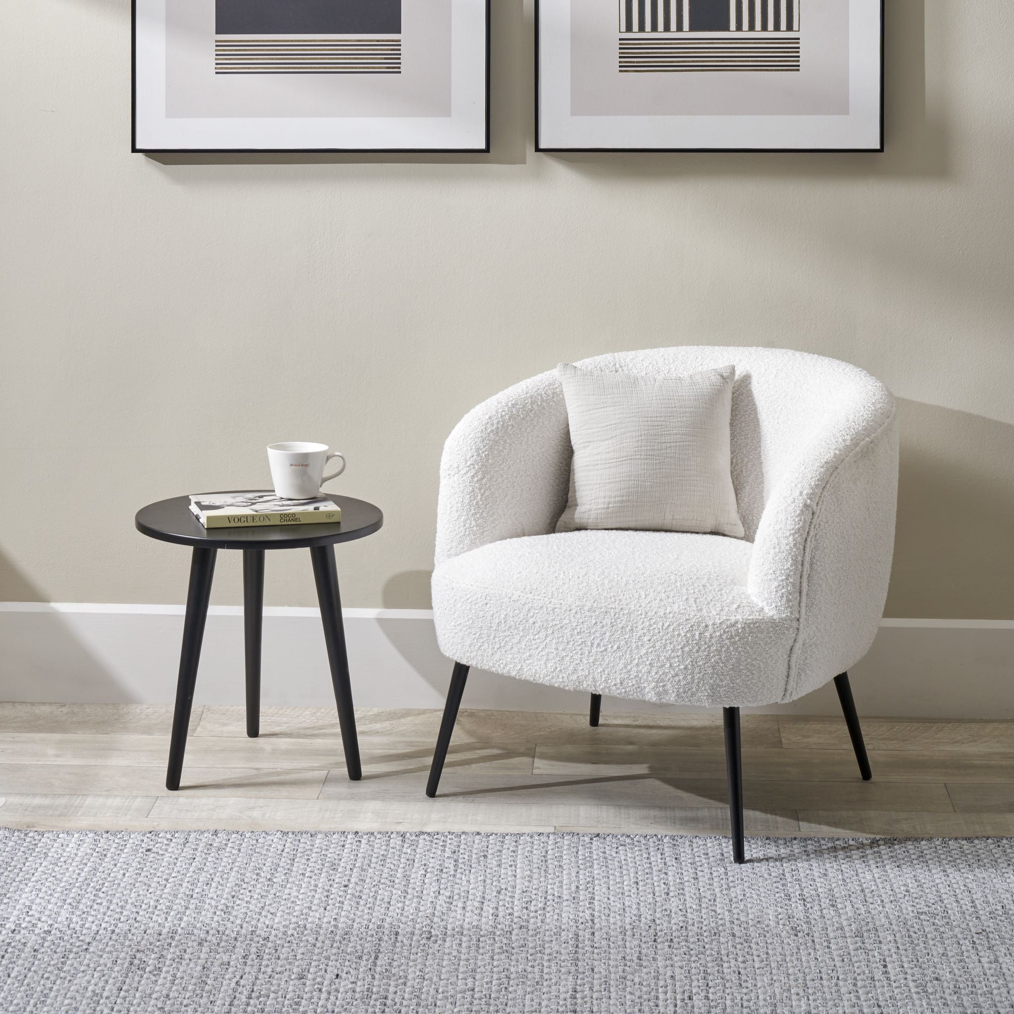 A Siena Bouclé Tub Chair with black legs from Pacific Lifestyle sits beside a small black round table holding a mug, with minimalist wall art adding a modern touch in the background.