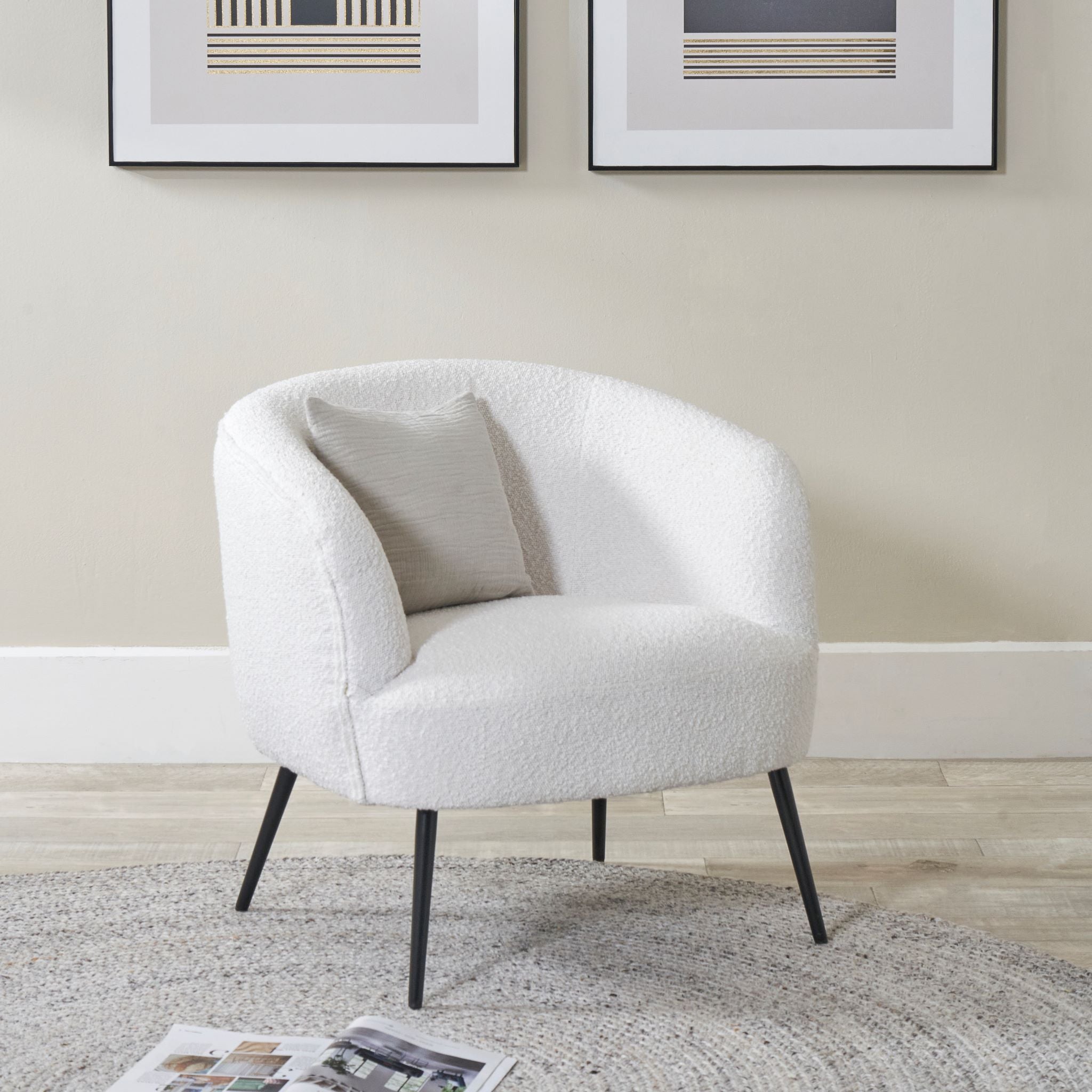 The Pacific Lifestyle Siena Bouclé Tub Chair With Black Legs, featuring modern white boucle upholstery and a light pillow, is elegantly placed in a room with framed art and a round rug on the wooden floor.