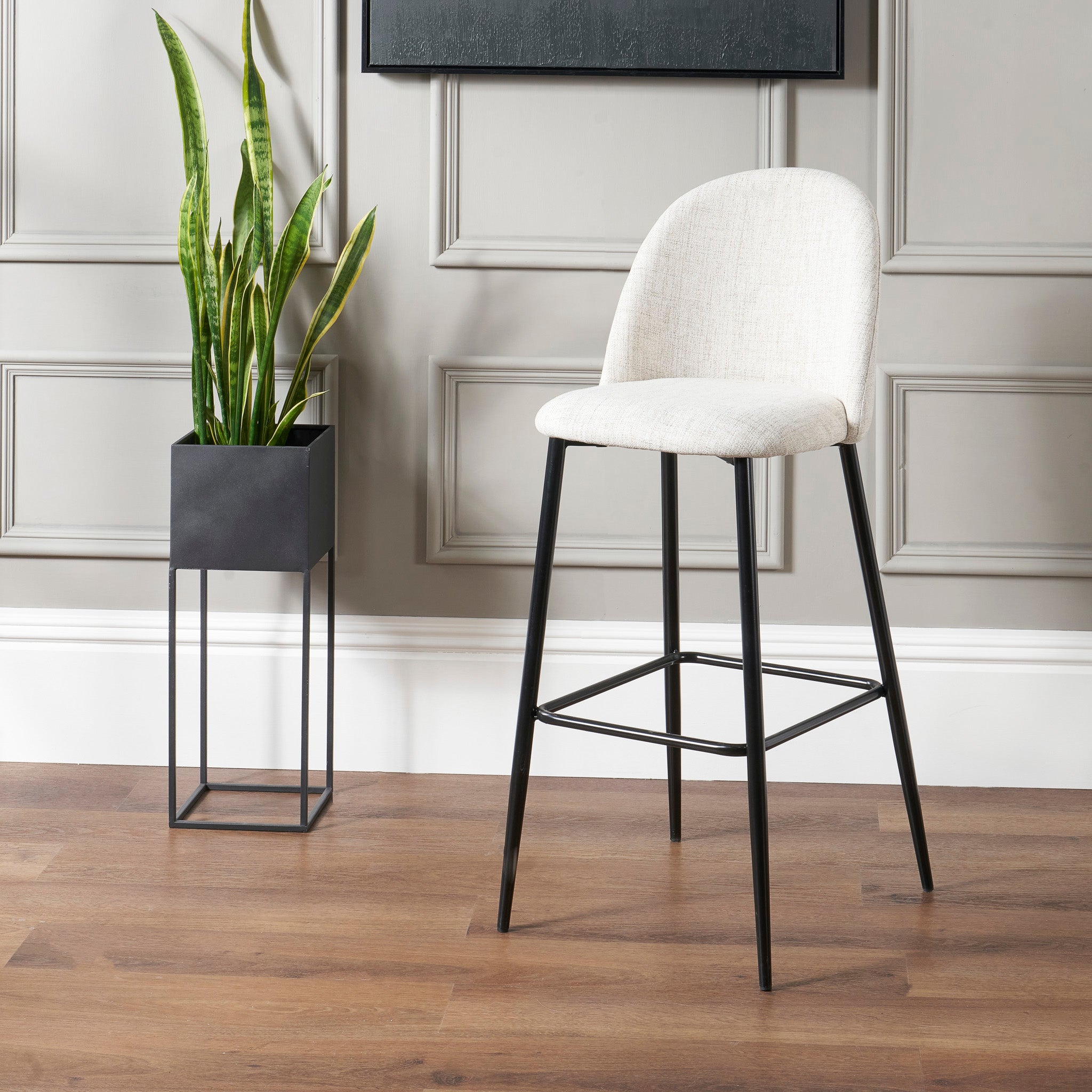 A contemporary Turi Pebble Linen Mix and Black Metal Bar Stool stands next to a tall potted plant on a wooden floor against a gray paneled wall.