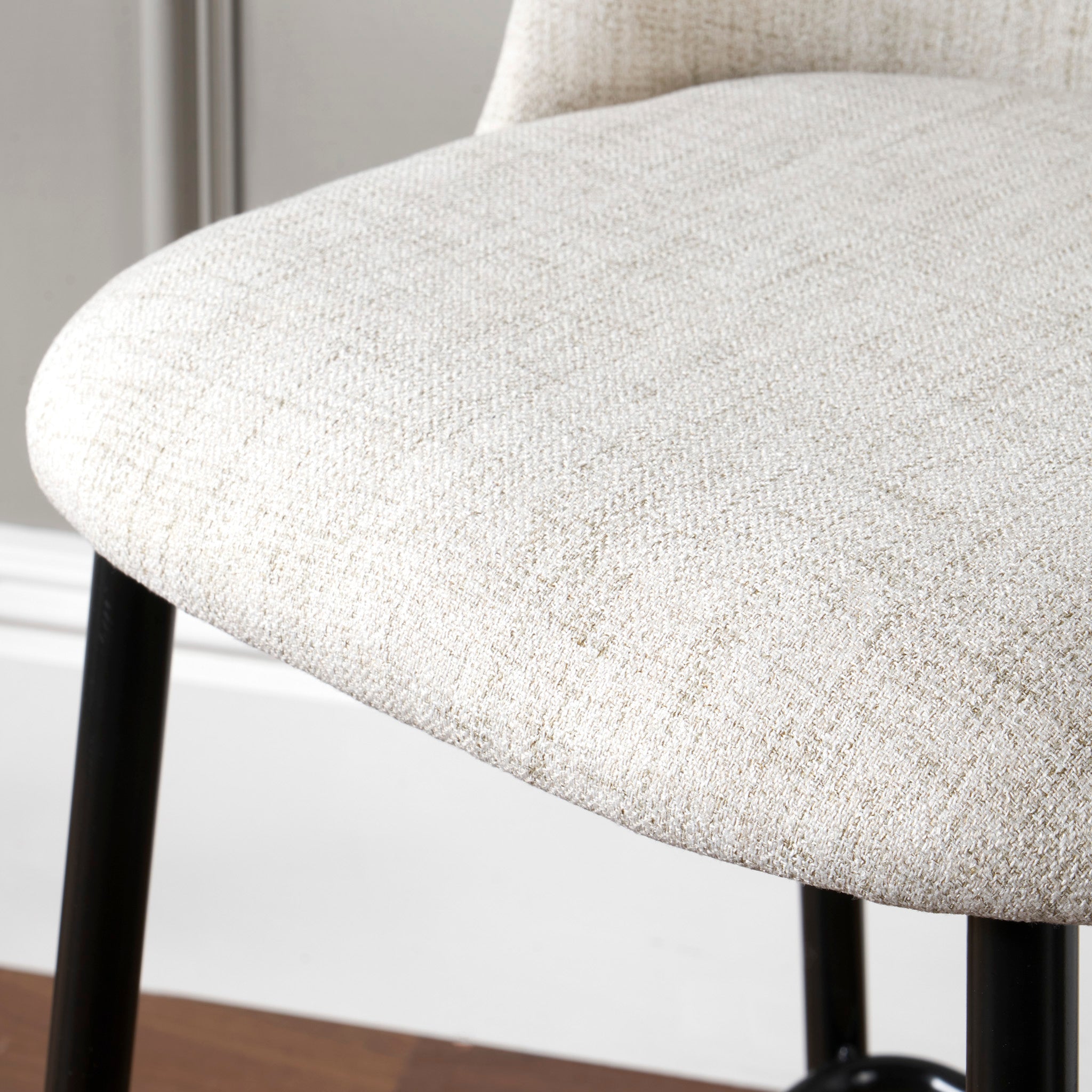 Close-up of a beige, upholstered Turi Pebble Linen Mix and Black Metal Bar Stool seat with black metal legs. The modern style chair is situated on a wooden floor.