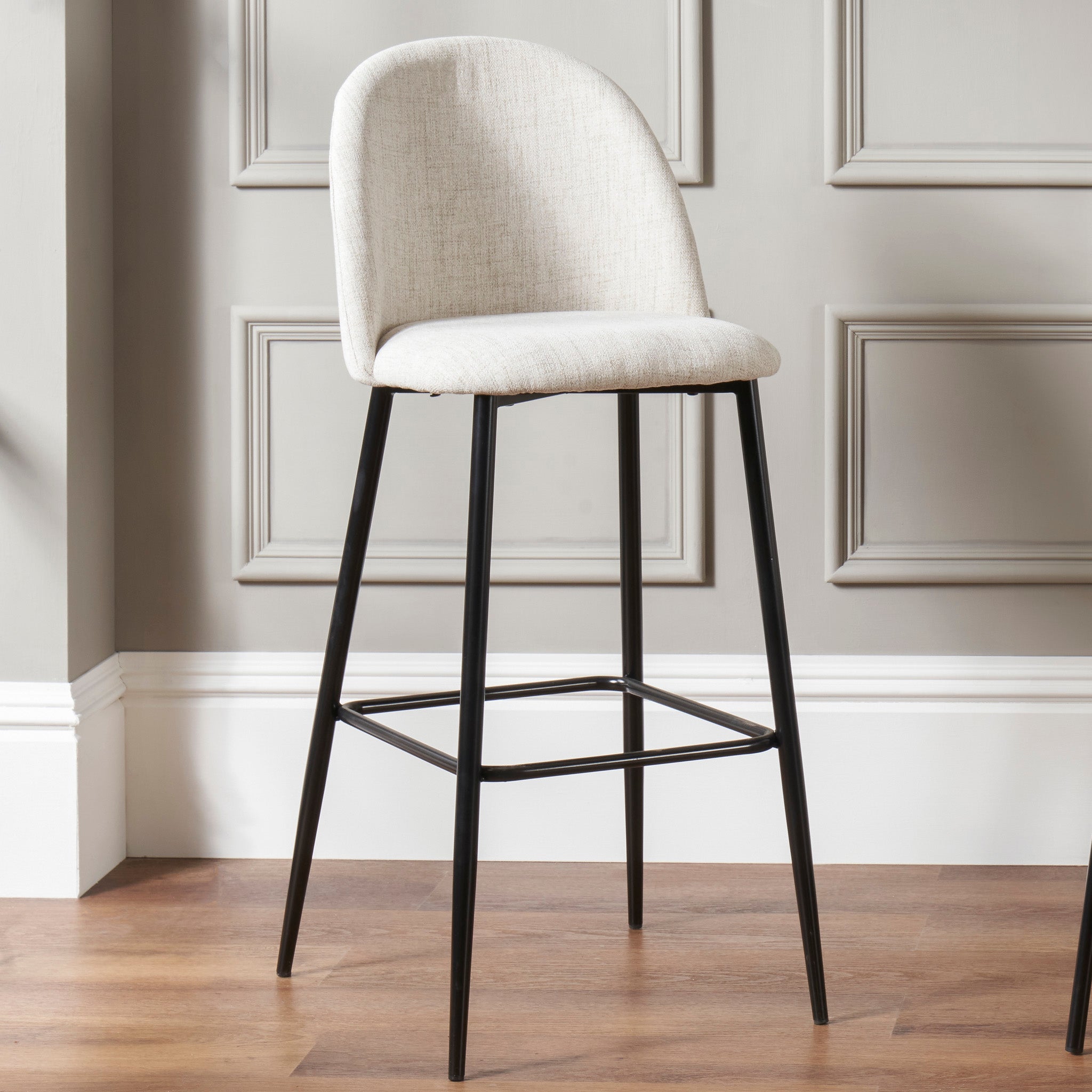 The Turi Pebble Linen Mix and Black Metal Bar Stool, a modern beige upholstered piece with a curved back and black metal legs, stands elegantly on the hardwood floor.