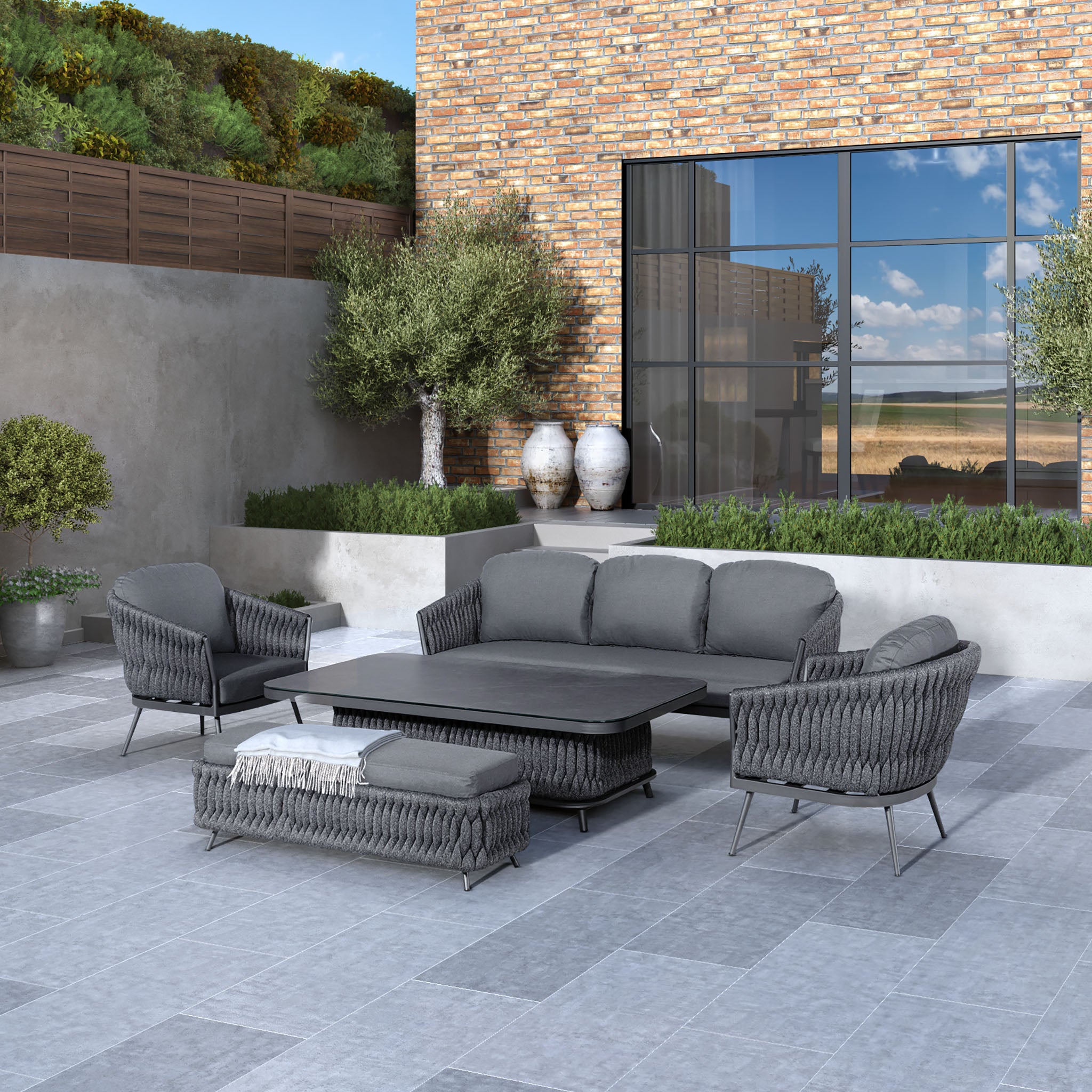 Modern patio with grey wicker furniture, including the UV-resistant Palma 3 Seat Rope Sofa Set with Rising Table in Grey, featuring a sofa, armchairs, and a sturdy aluminium coffee table, all set against a brick wall.