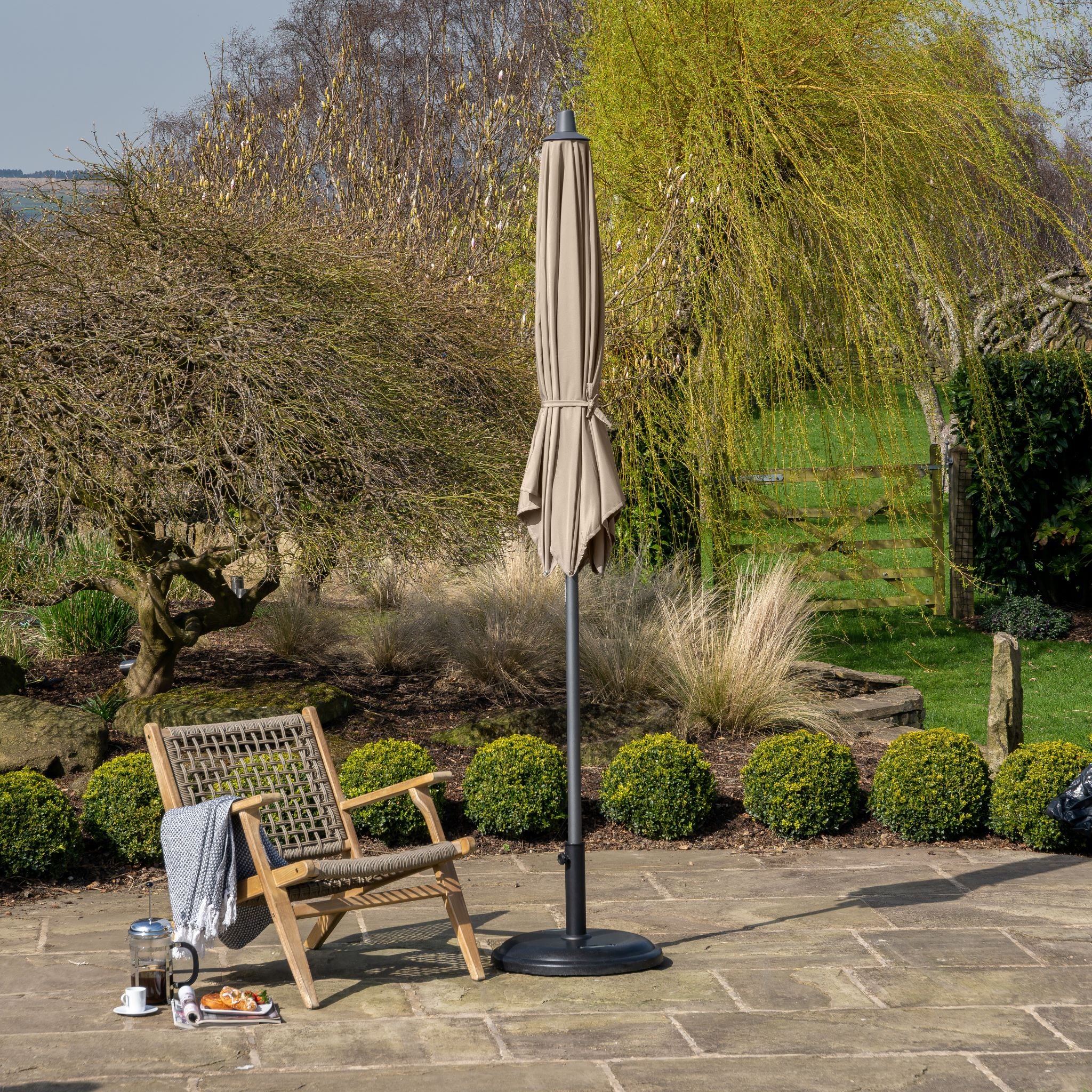 A wooden chair and the Pacific Lifestyle - Riva 2.5m Round Taupe Parasol on a stone patio, surrounded by lush greenery and bushes.