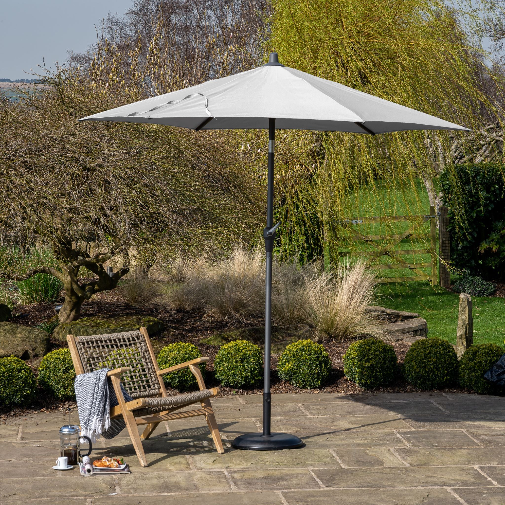 A patio scene features a wooden chair with a blue cushion, complemented by a coffee set under the expansive Pacific Lifestyle - Riva 3m Round Grey Parasol with UV protection. Surrounded by lush greenery, the parasol's powder-coated aluminium frame adds elegance to the setting.