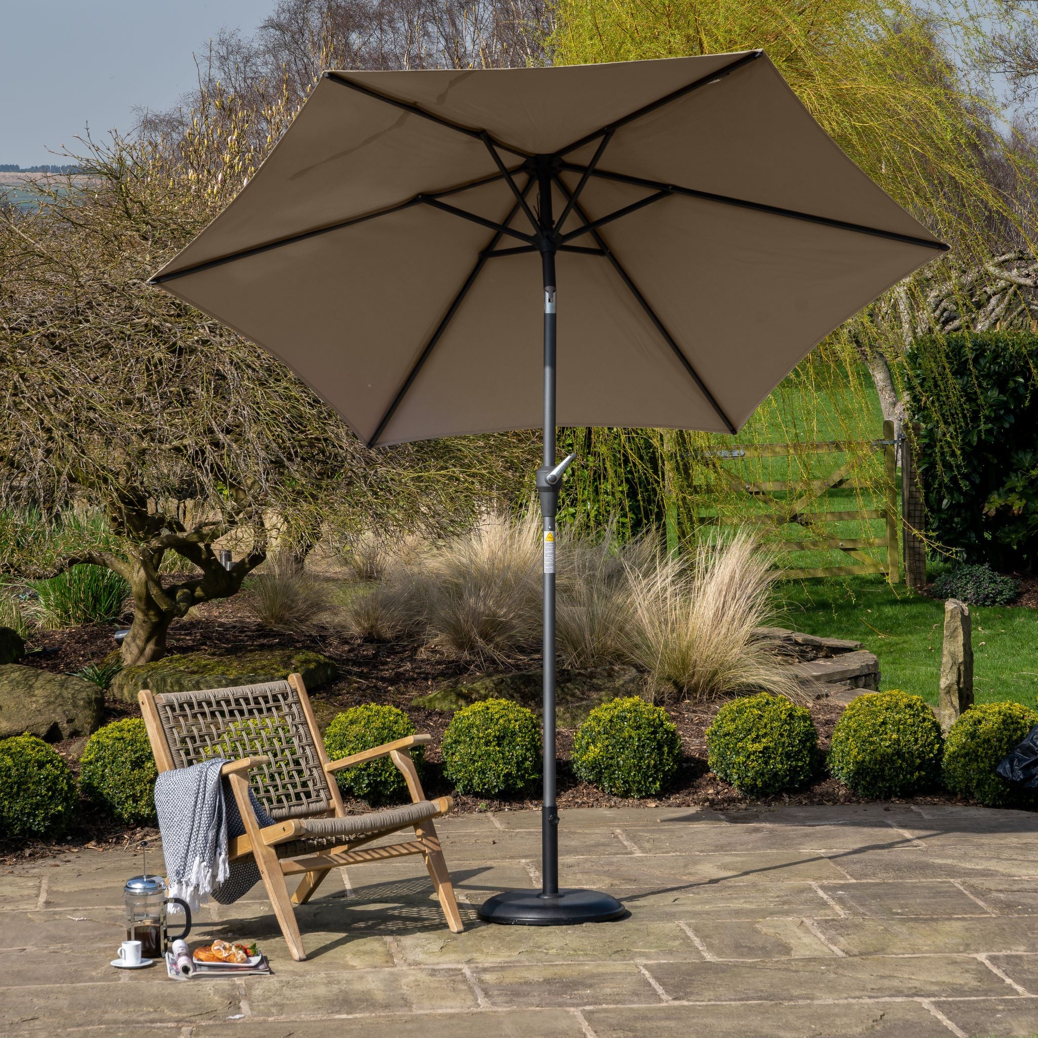 Patio with a wooden chair, a small table with a coffee setup, and a Riva 3m Round Taupe Parasol featuring a premium-class fabric canopy in a lush garden setting.