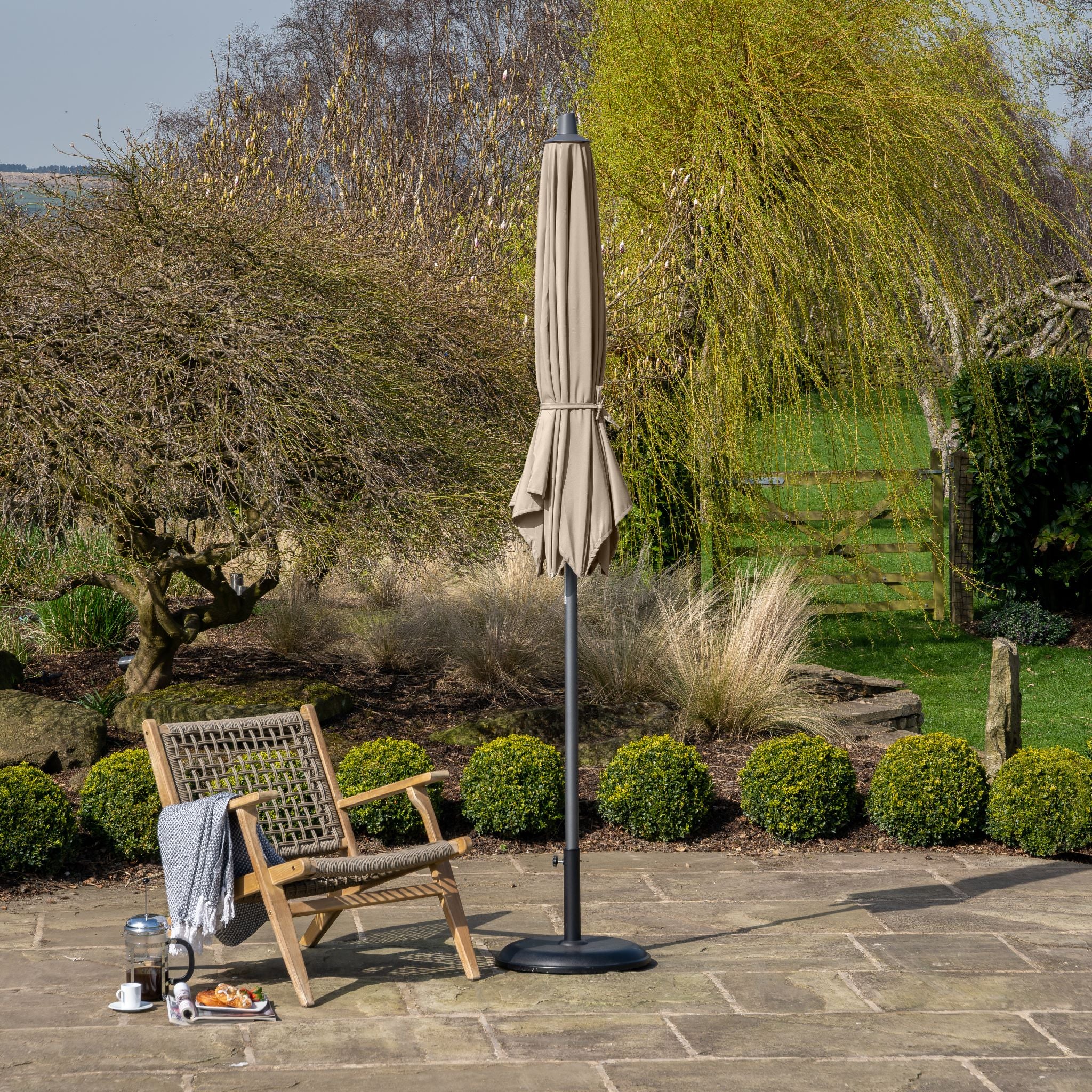 The outdoor patio scene features a wooden chair and a Riva 3m Round Taupe Parasol with a premium-class fabric canopy, along with a blanket and a tray holding coffee on the stone-tiled surface.