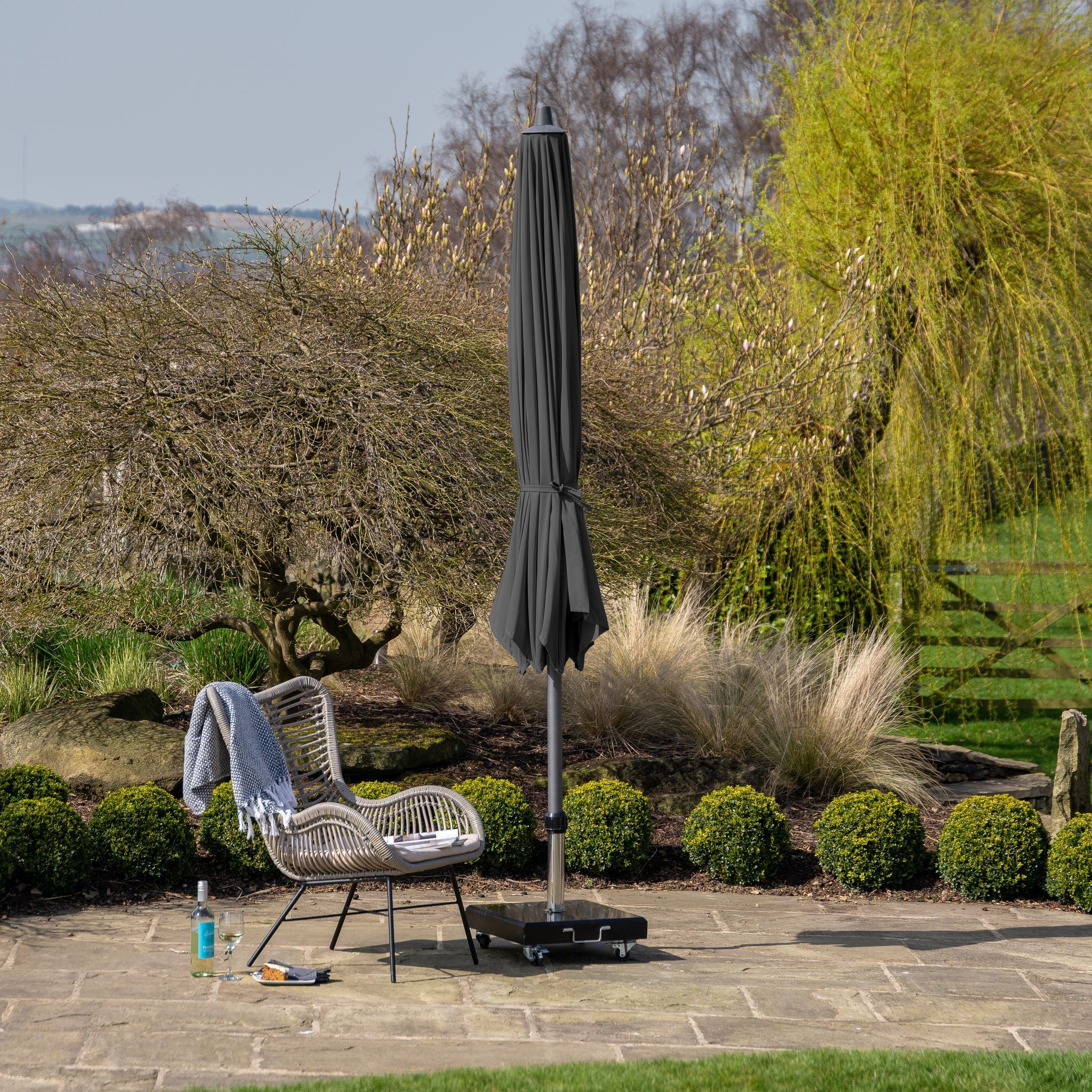 A patio featuring a wicker chair, the folded Riva 3.5m Round Anthracite Parasol, a drink bottle, and a book, set in a garden with trees and bushes in the background. The setup highlights premium-class fabric upholstery and durable powder-coated aluminium frames.