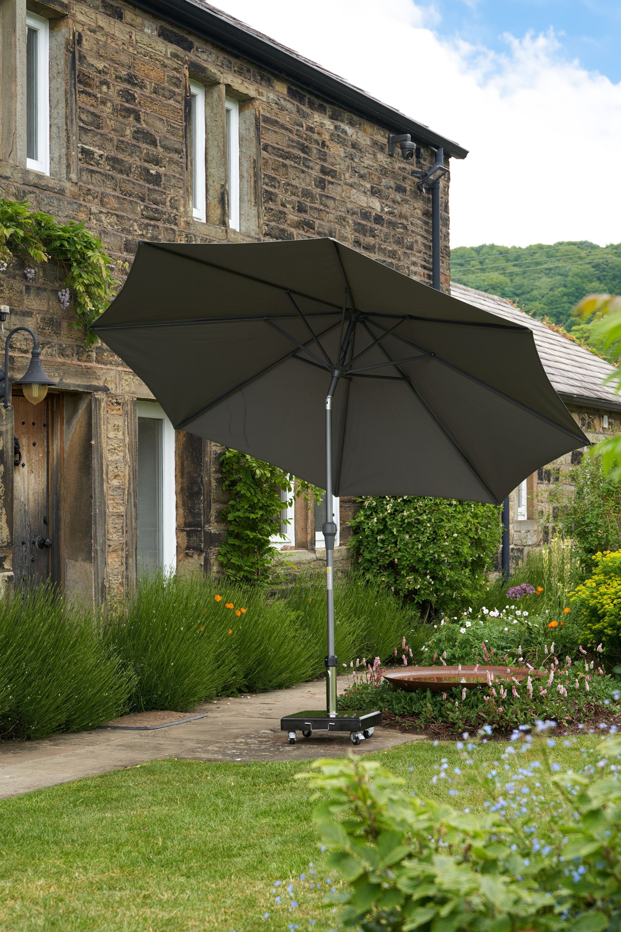 The Pacific Lifestyle Riva 3.5m Round Anthracite Parasol, with an aluminum frame, stands open in a garden outside a stone house, providing UV protection amidst the lush greenery and vibrant flowers.