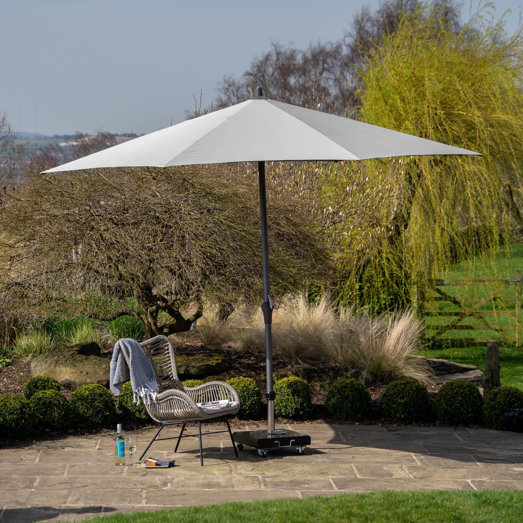 Outdoor patio with a Riva 3.5m Round Grey Parasol providing UV protection over a wicker chair, a gray blanket on the chair, and a drink bottle on the ground.