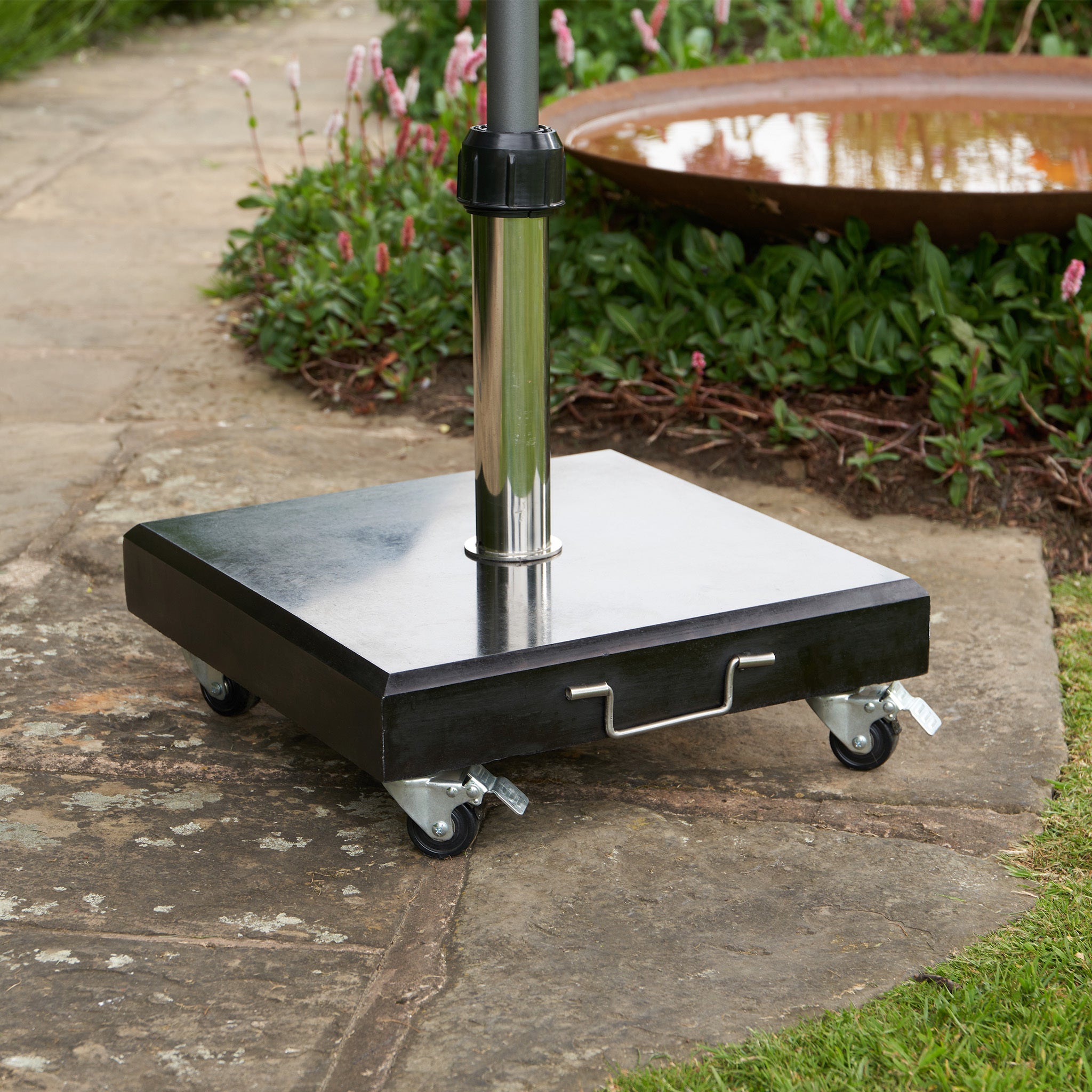 A Rome Polished Black Granite 40KG Parasol Base on wheels, positioned on a stone patio with greenery and a water feature in the background.