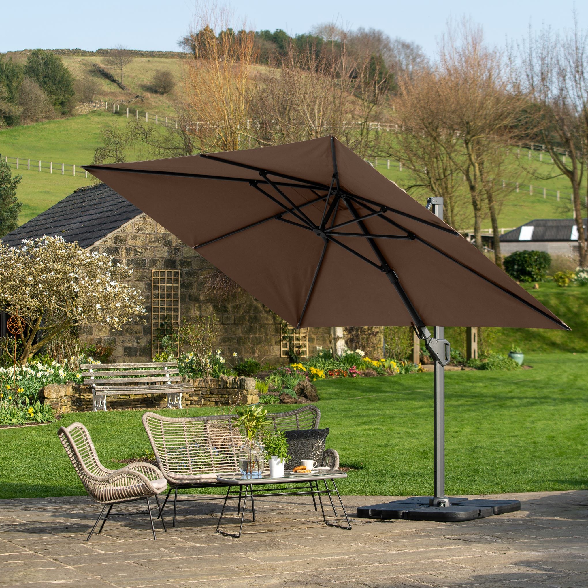 The patio features wicker seating and a table under the large Pacific Lifestyle Challenger T2 3.5m x 2.6m Rectangular Cantilever Parasol in Taupe with tilt parasol technology for optimal UV protection, set against a backdrop of garden and rolling hills.