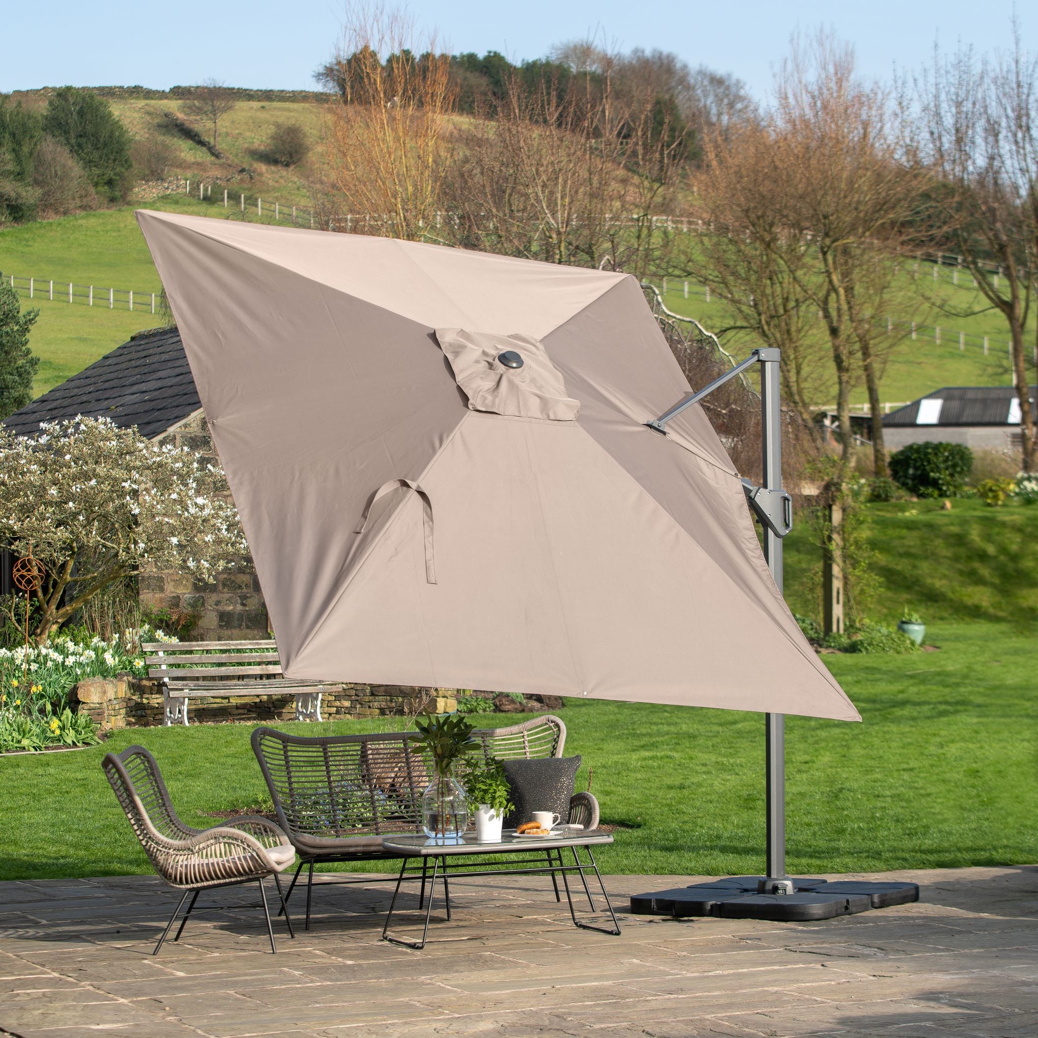 The Pacific Lifestyle Challenger T2 3.5m x 2.6m Rectangular Cantilever Parasol in Taupe offers UV protection, shading a wicker table set on a stone patio against a grassy background, while its sturdy, powder-coated aluminium frame ensures durability and style.