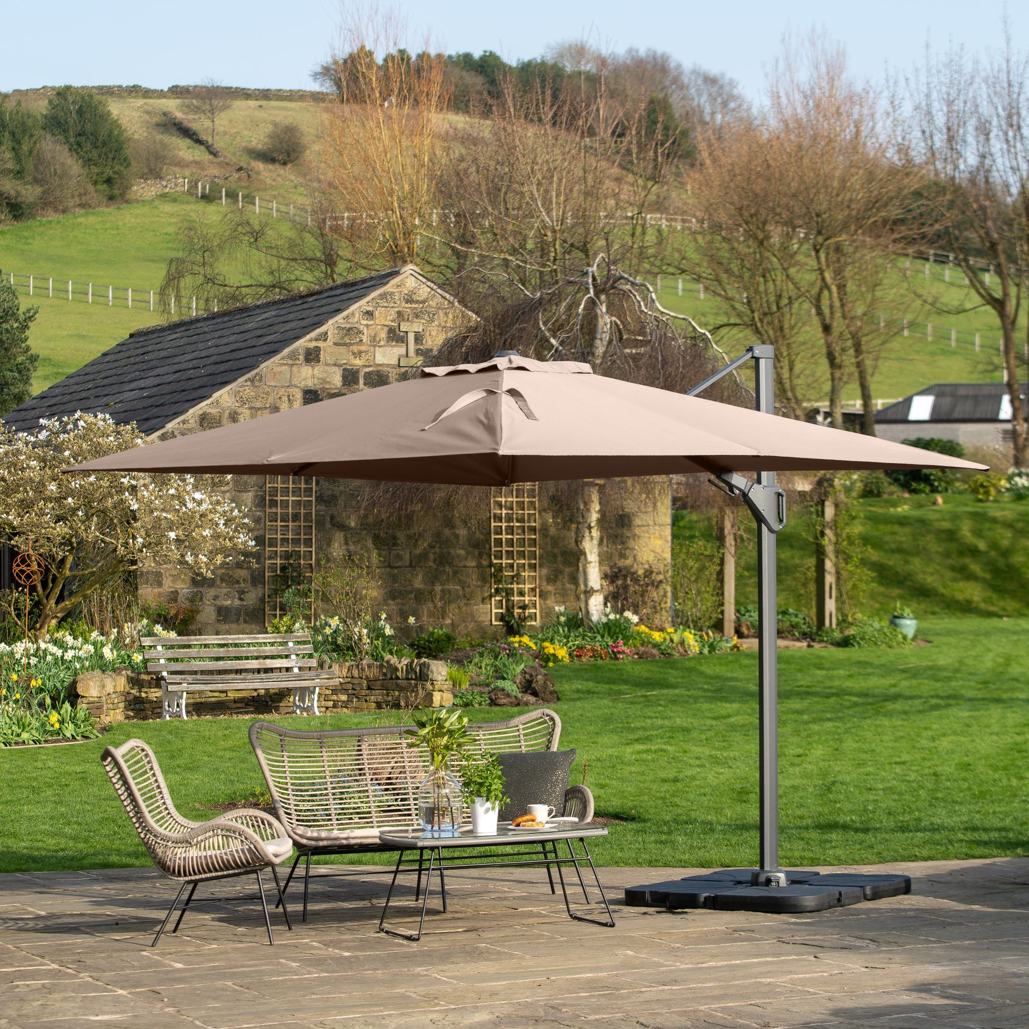 A charming patio showcases wicker furniture under the Pacific Lifestyle Challenger T2 Rectangular Cantilever Parasol in Taupe, offering UV protection, nestled beside a garden and rustic stone building.