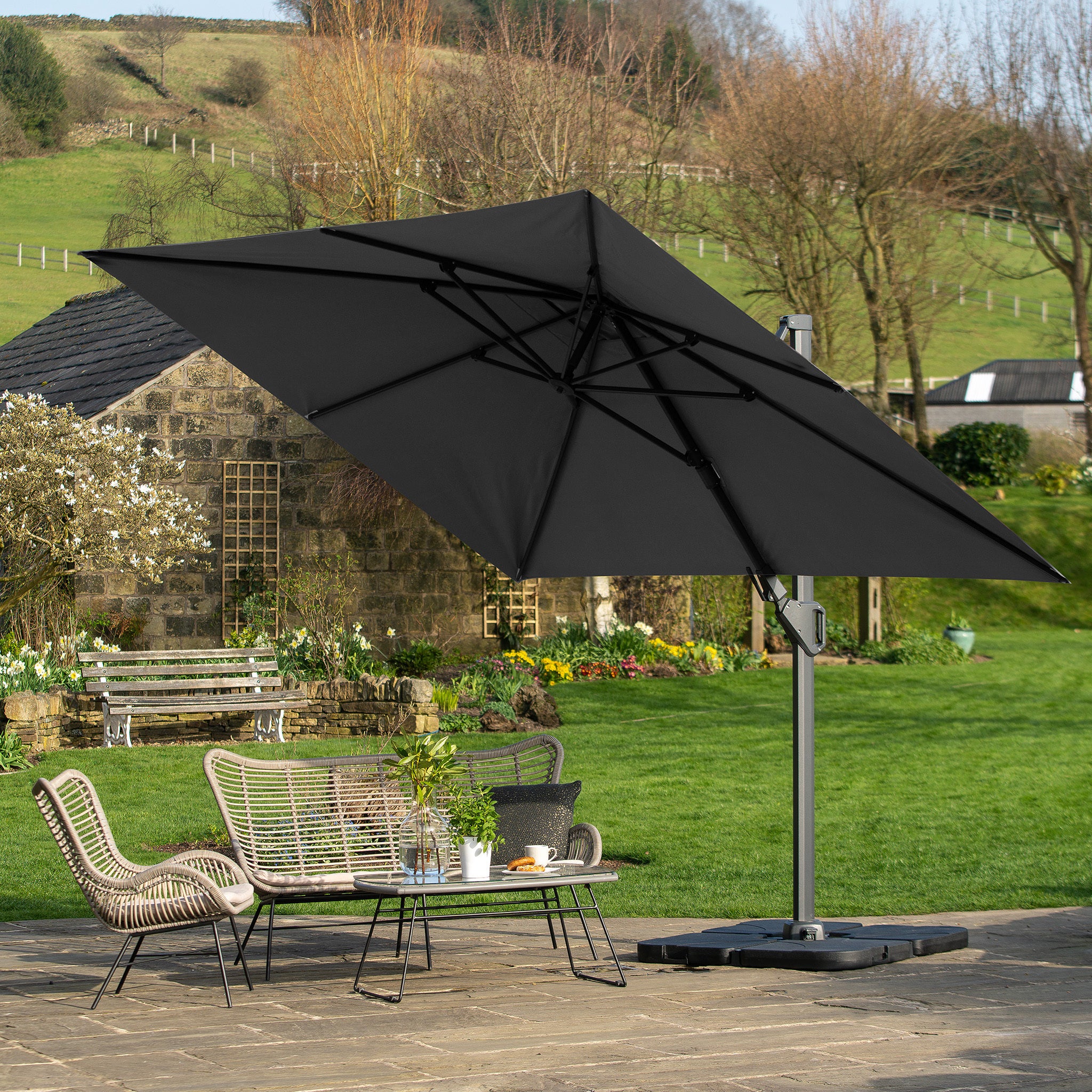 A stone terrace with a Pacific Lifestyle Challenger T2 3.5m x 2.6m Rectangular Cantilever Parasol in Anthracite overlooks a lush lawn and rustic building, featuring a double tilt system and premium-class fabric for style and durability.