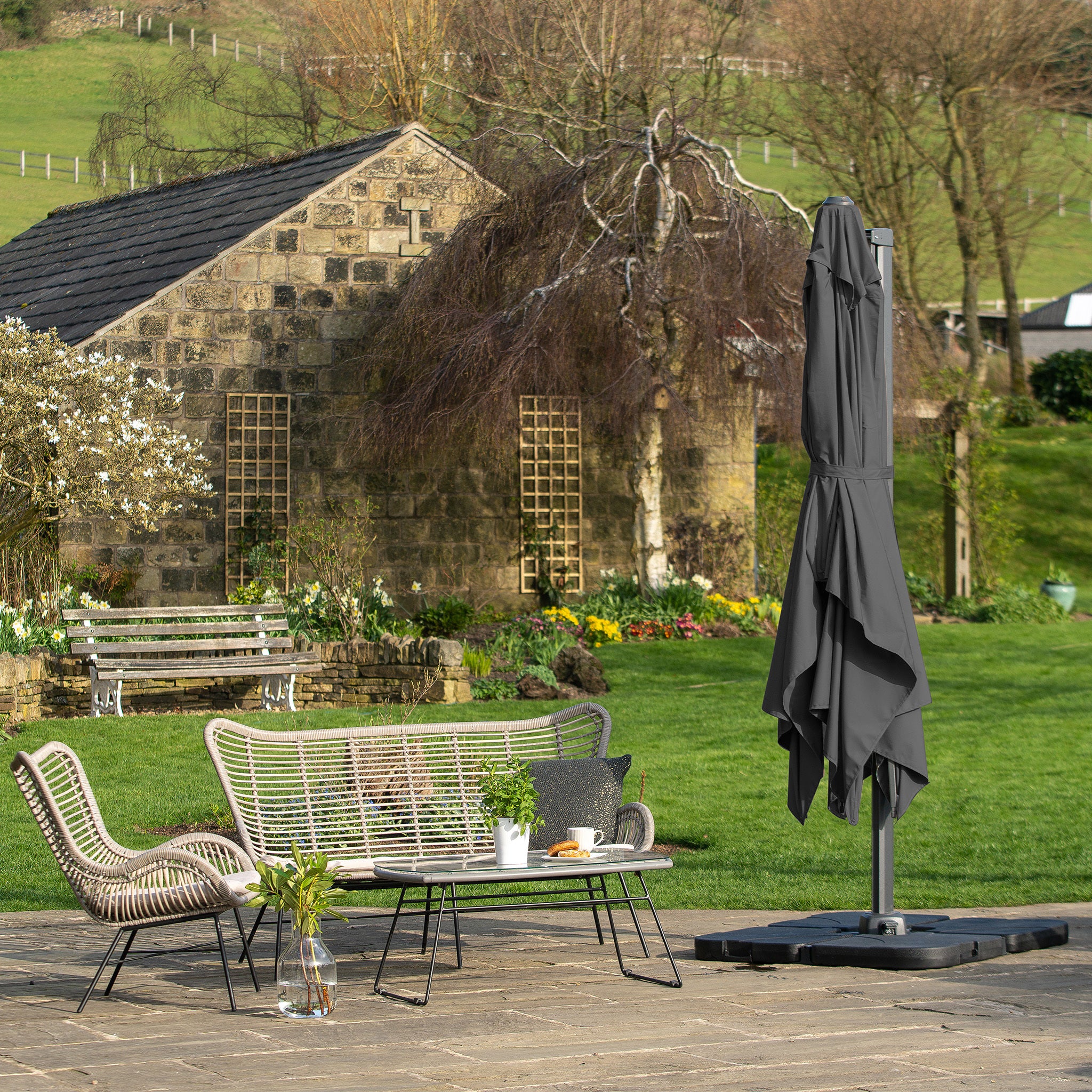 A cozy patio features wicker furniture and a folded Pacific Lifestyle Challenger T2 3.5m x 2.6m Rectangular Cantilever Parasol in Anthracite, with premium fabric, nestled amid greenery and a rustic stone building.