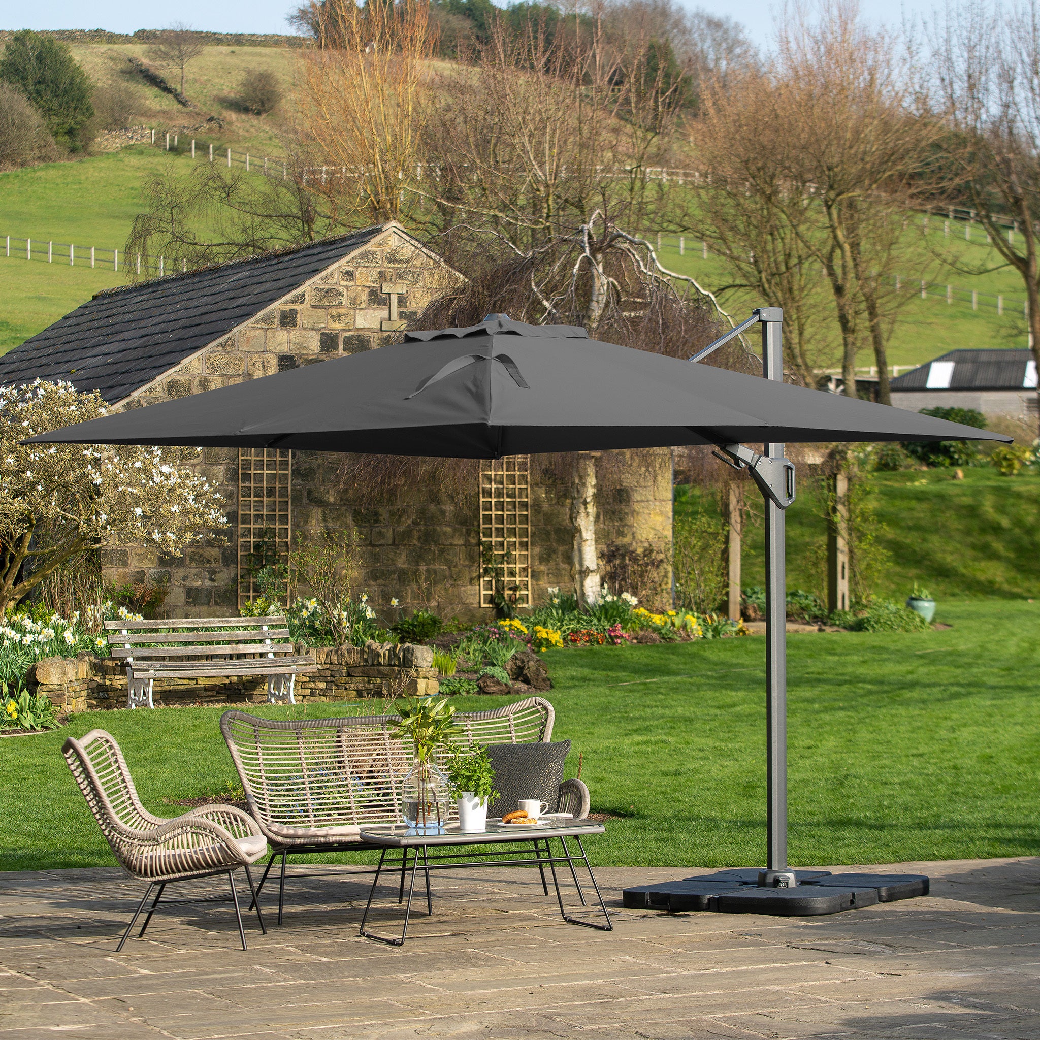 A patio scene features the Pacific Lifestyle Challenger T2 3.5m x 2.6m Rectangular Cantilever Parasol in Anthracite, its dual tilt system shading wicker chairs and a table. The premium fabric exudes elegance against a garden and stone building backdrop.