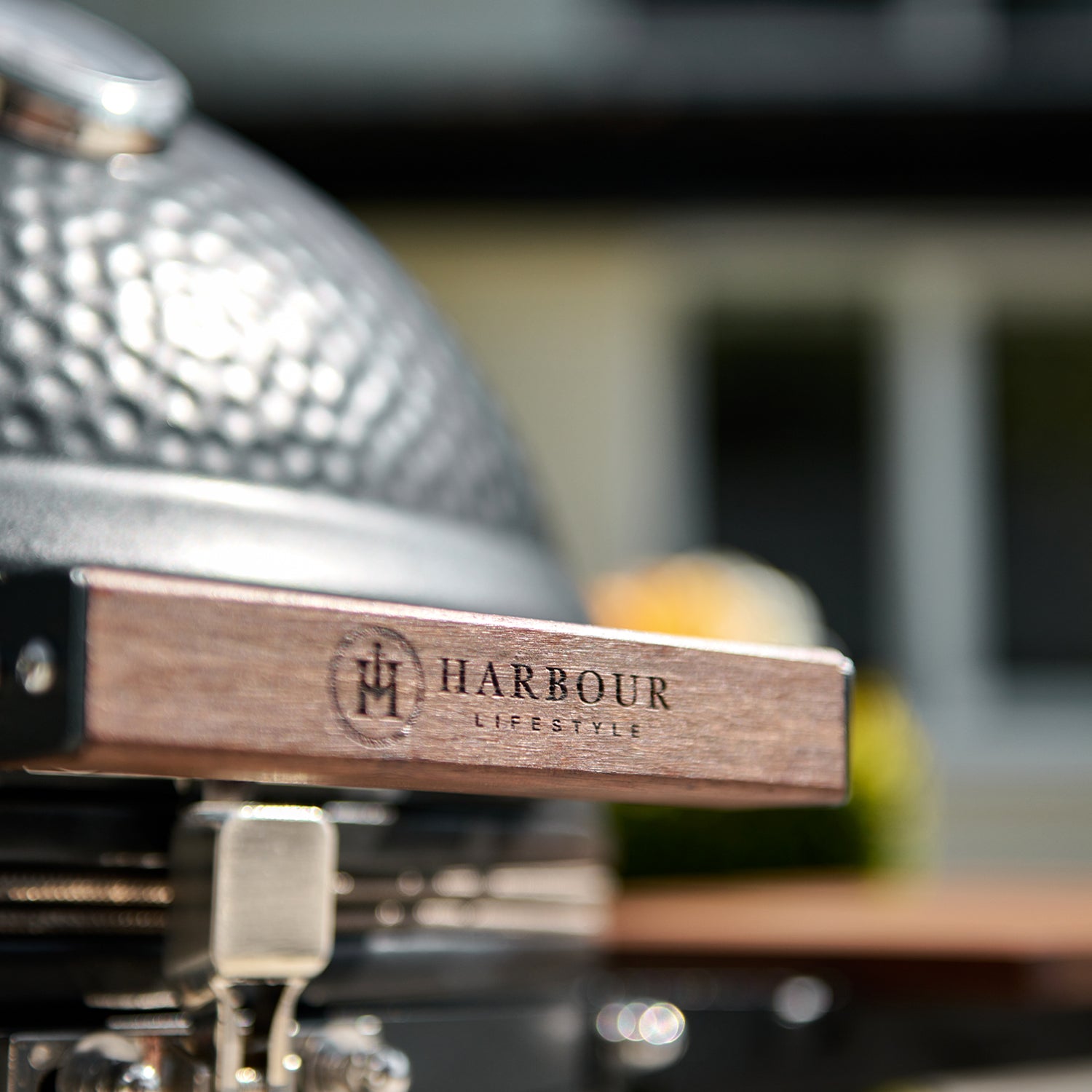 Close-up of a barbecue grill handle with the brand "Harbour Lifestyle" engraved on it. The Kamado BBQ 22" Ceramic Grill in Black, known for its exceptional ceramic design, adds a touch of elegance in the blurred background.