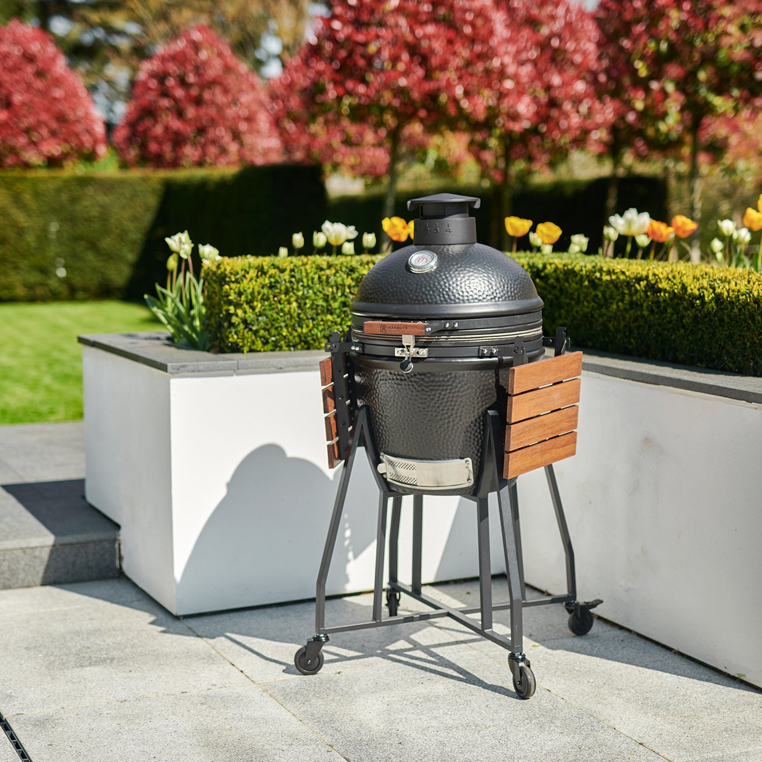 A Kamado BBQ 22" Ceramic Grill in Black, complete with wooden side shelves, sits on a patio surrounded by greenery and blooming flowers.