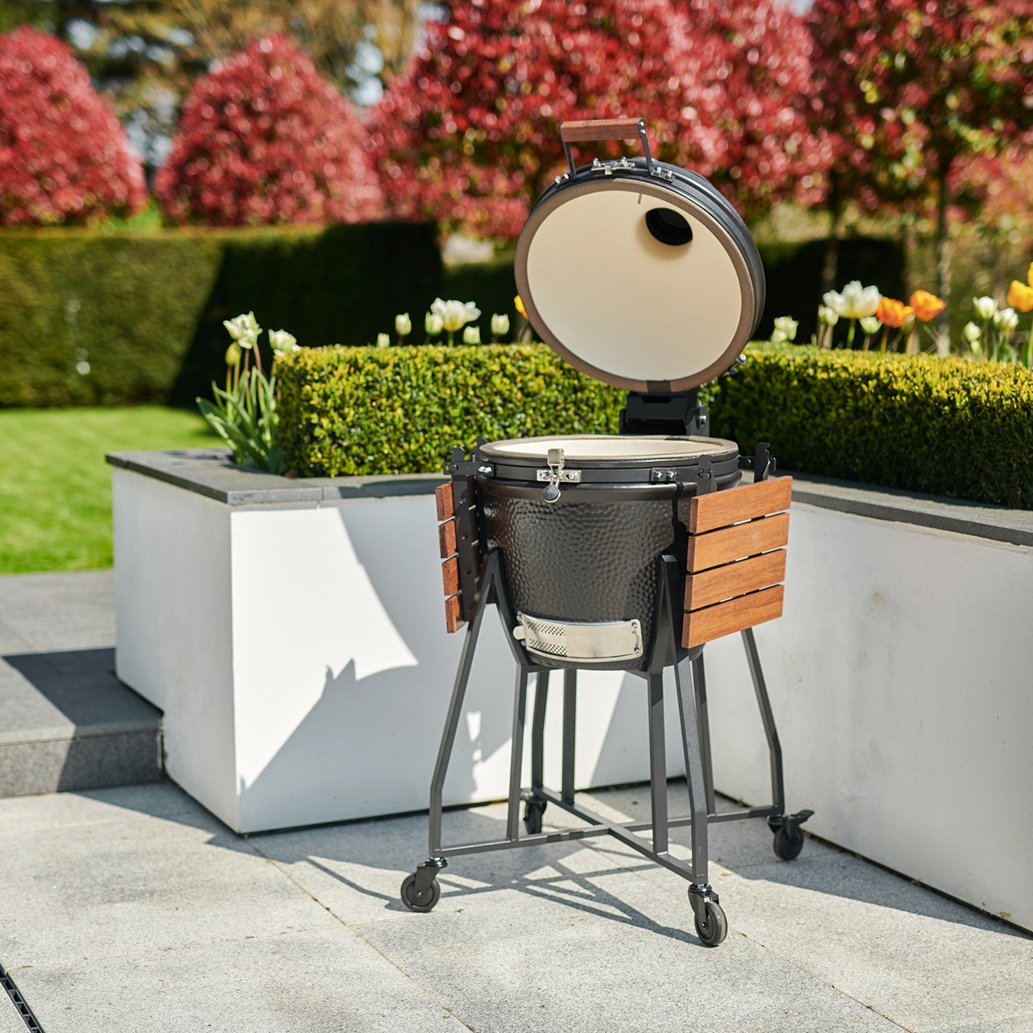 A Kamado BBQ 22" Ceramic Grill in Black, featuring a wooden side shelf, stands open on the patio, with a garden and vibrant flowers in the background.