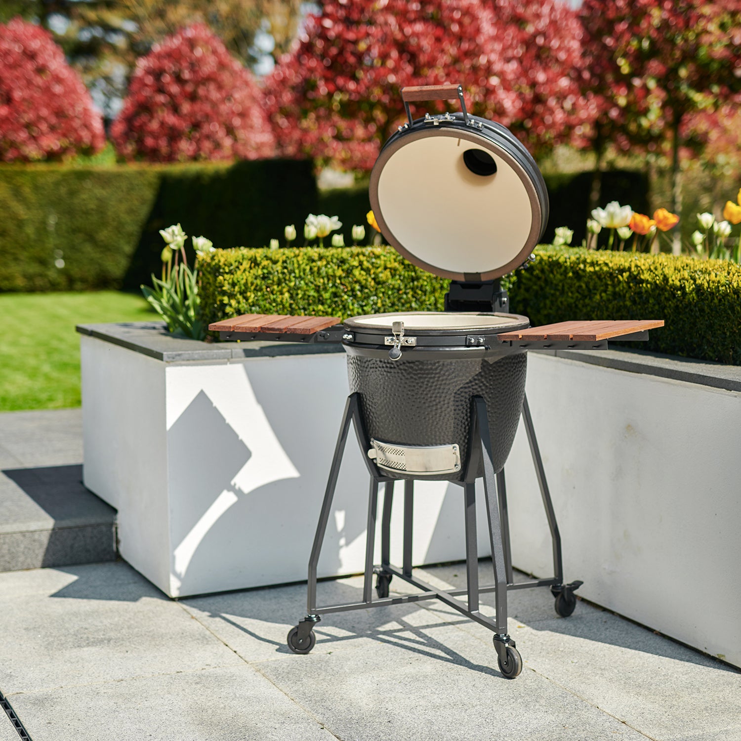 A Kamado BBQ 22" Ceramic Grill in black, with an open lid, set on a patio next to a garden with blooming flowers and trimmed hedges.