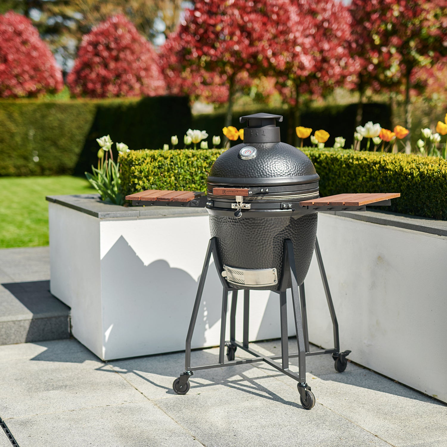 A Kamado BBQ 22" Ceramic Grill in Black, complete with wooden side tables, is set up in a neatly landscaped garden with colorful flowers and trimmed hedges.