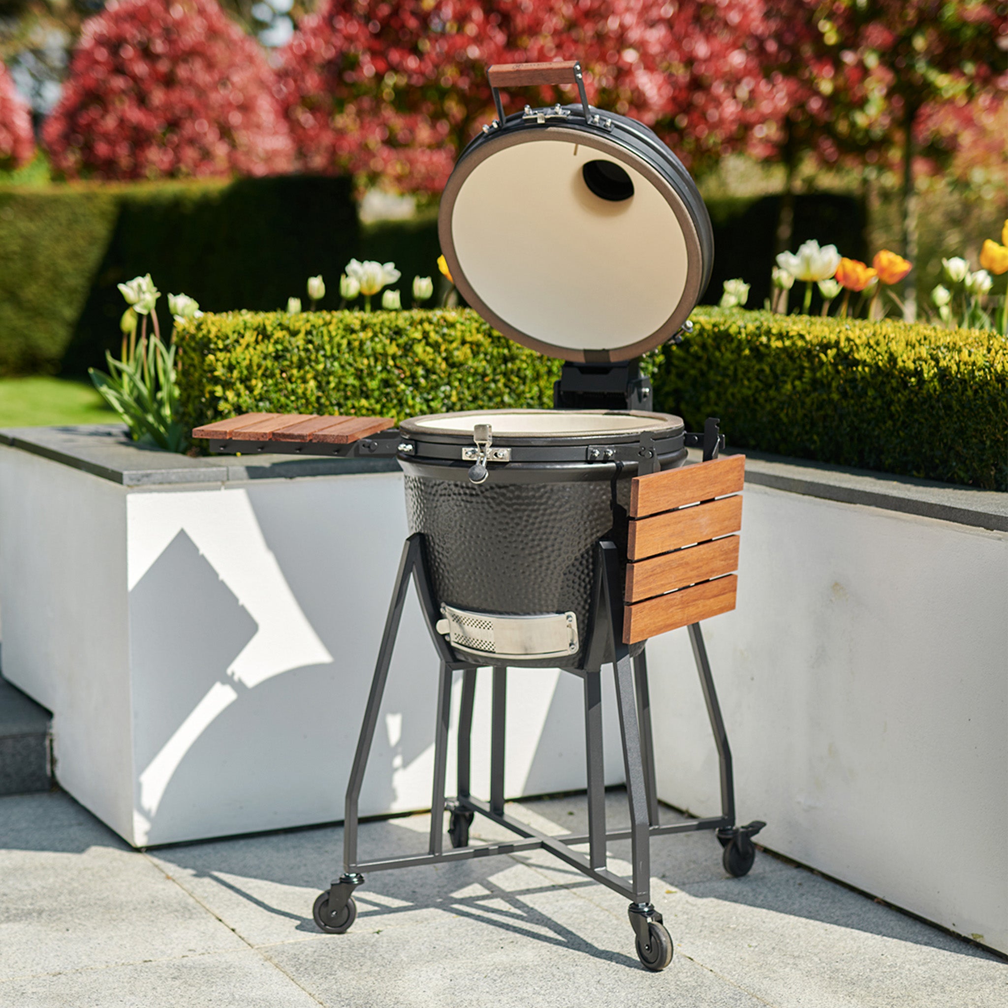 A Kamado BBQ 22" Ceramic Grill in Black with a hinged lid stands on a patio, surrounded by greenery and blooming tulips.