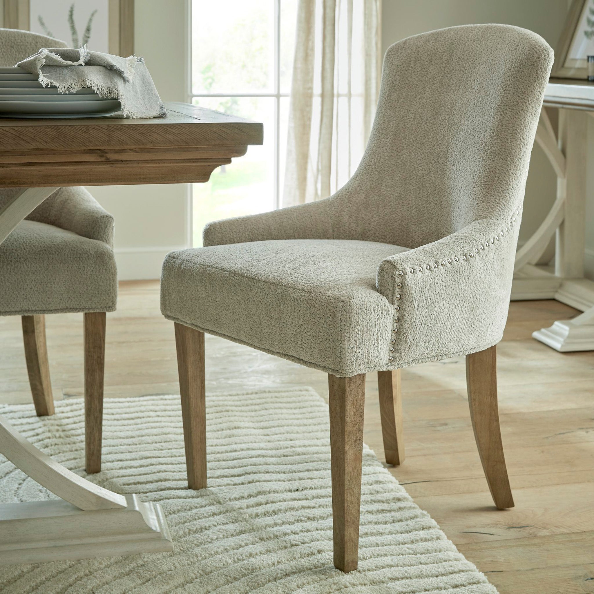 A Brockham Taupe Dining Chair crafted with taupe woven fabric is elegantly placed on a light wooden floor near a table, complemented by sheer curtains in the backdrop. Its rubberwood frame enhances the room's sophisticated atmosphere.