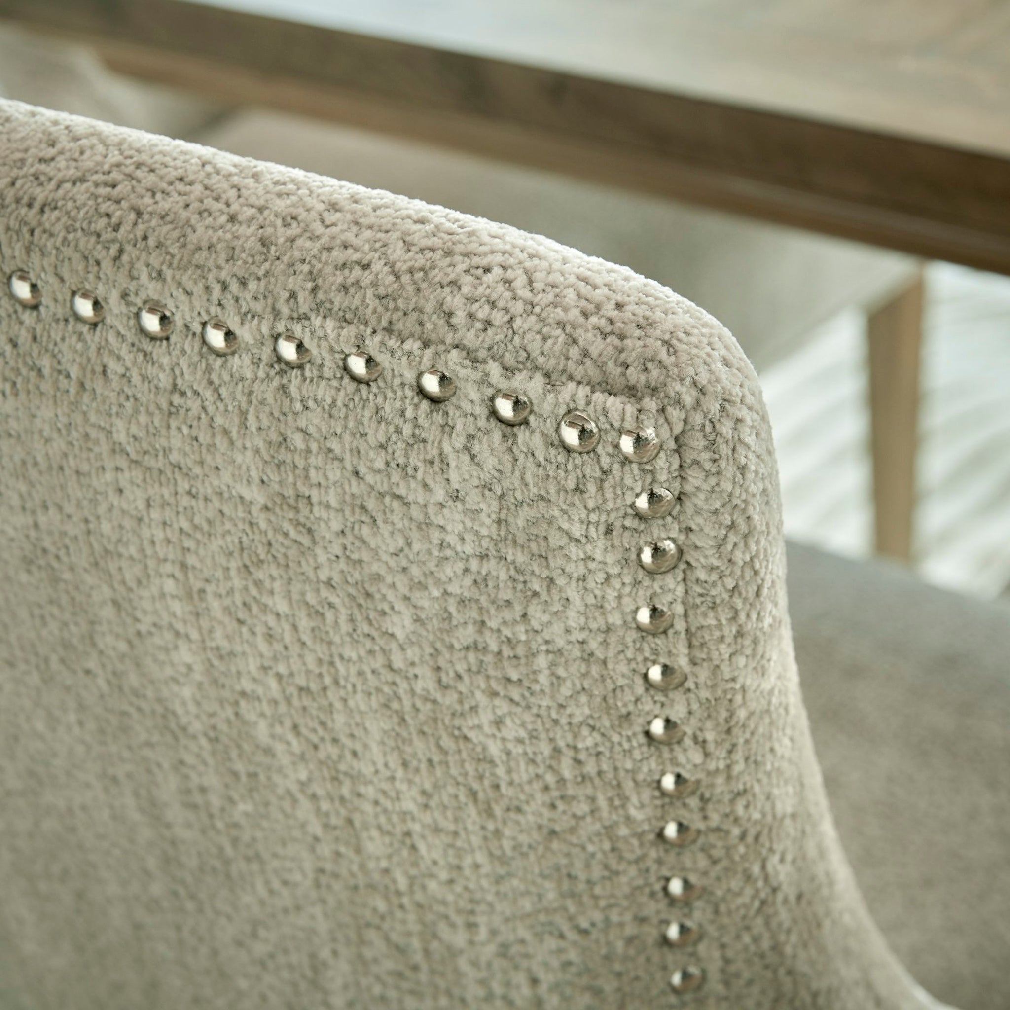 A detailed view of the Brockham Taupe Dining Chair, which has a rubberwood frame and a textured back embellished with metal studs along the edge, placed next to a wooden table.