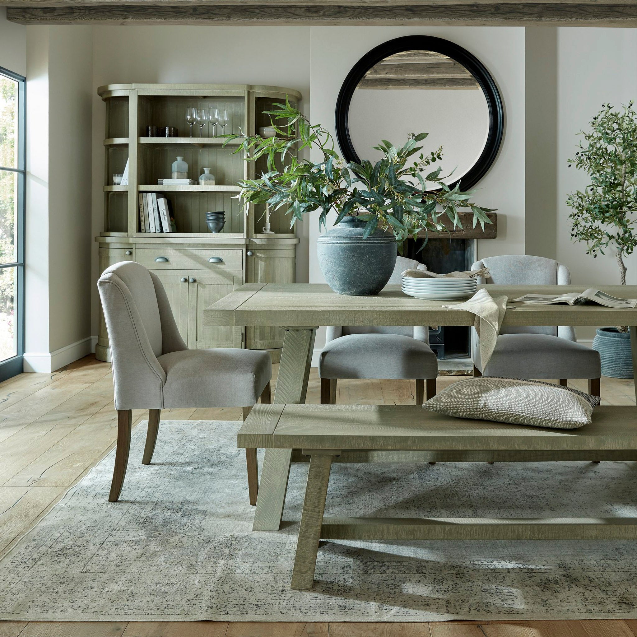 A rustic dining room showcases a wooden table accompanied by Kingham Grey Dining Chairs and a bench, all enhanced by a potted plant and a circular mirror on the wall. The touch of rubberwood in the furniture adds an extra layer of charm to the cozy setup.