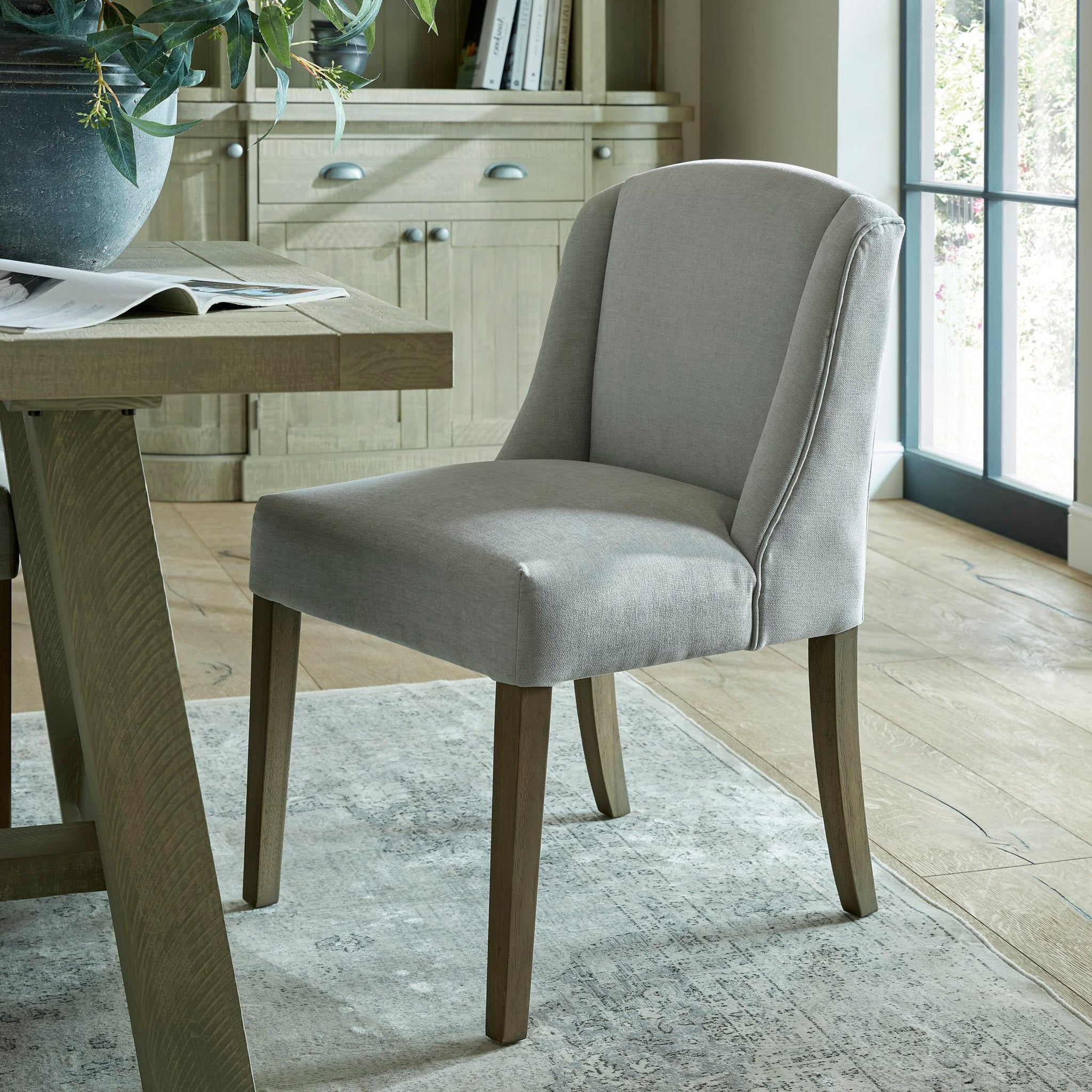 The Kingham Grey Dining Chair, featuring woven fabric, sits next to a wooden dining table in a sunlit room, complete with a rug and cabinet.