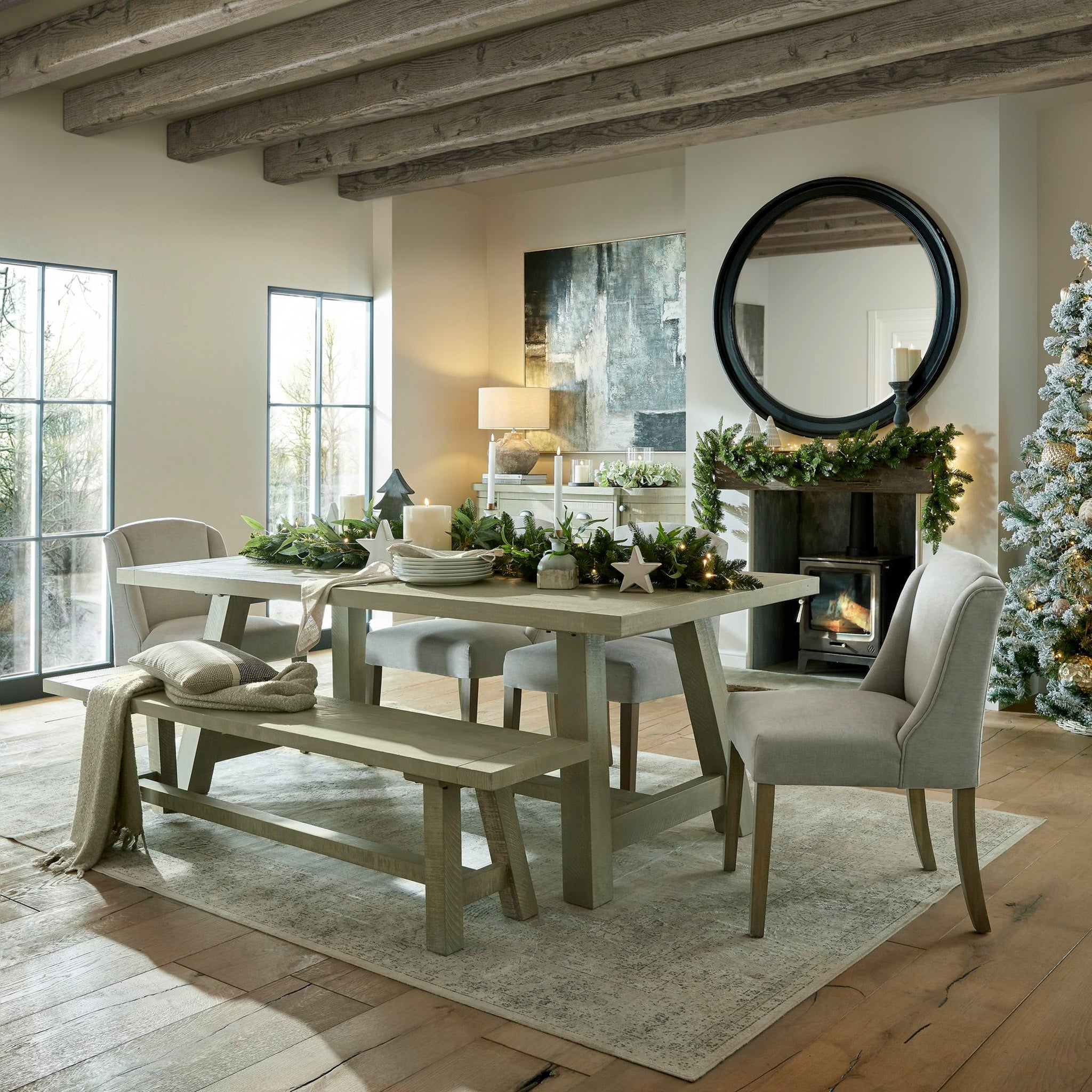 A cozy dining room with rustic decor showcases a festive table setting, highlighted by the Kingham Grey Dining Chairs in their light grey woven fabric. In the corner stands a Christmas tree adorned with twinkling lights, while candles and garlands contribute to the warm glow of the space.