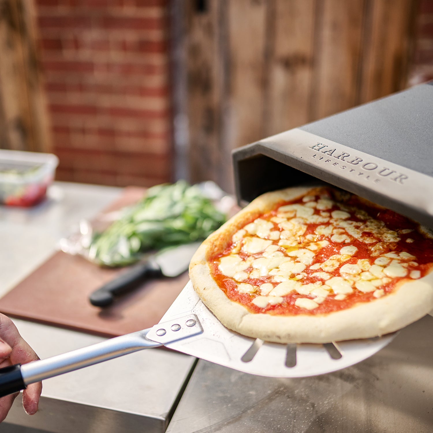 A hand uses the Juno 12" Pizza Oven Bundle with Gas Burner to remove a fresh Margherita pizza, showcasing its stainless steel construction.