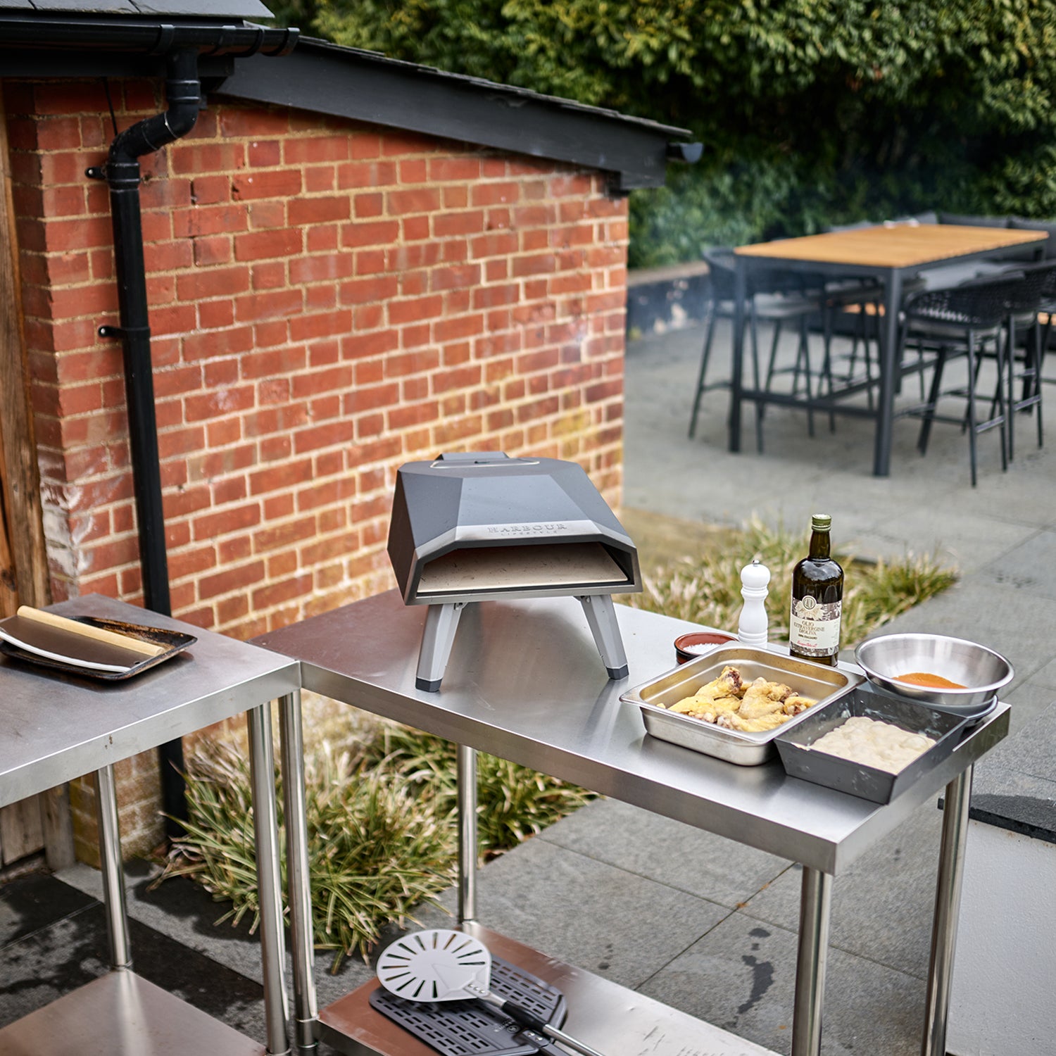 Outdoor setup featuring the Juno 12" Pizza Oven Bundle with Gas Burner on a table beside cooking ingredients, near a brick wall and a dining area. The stainless steel construction ensures durability, promising delicious stone-baked pizza every time.