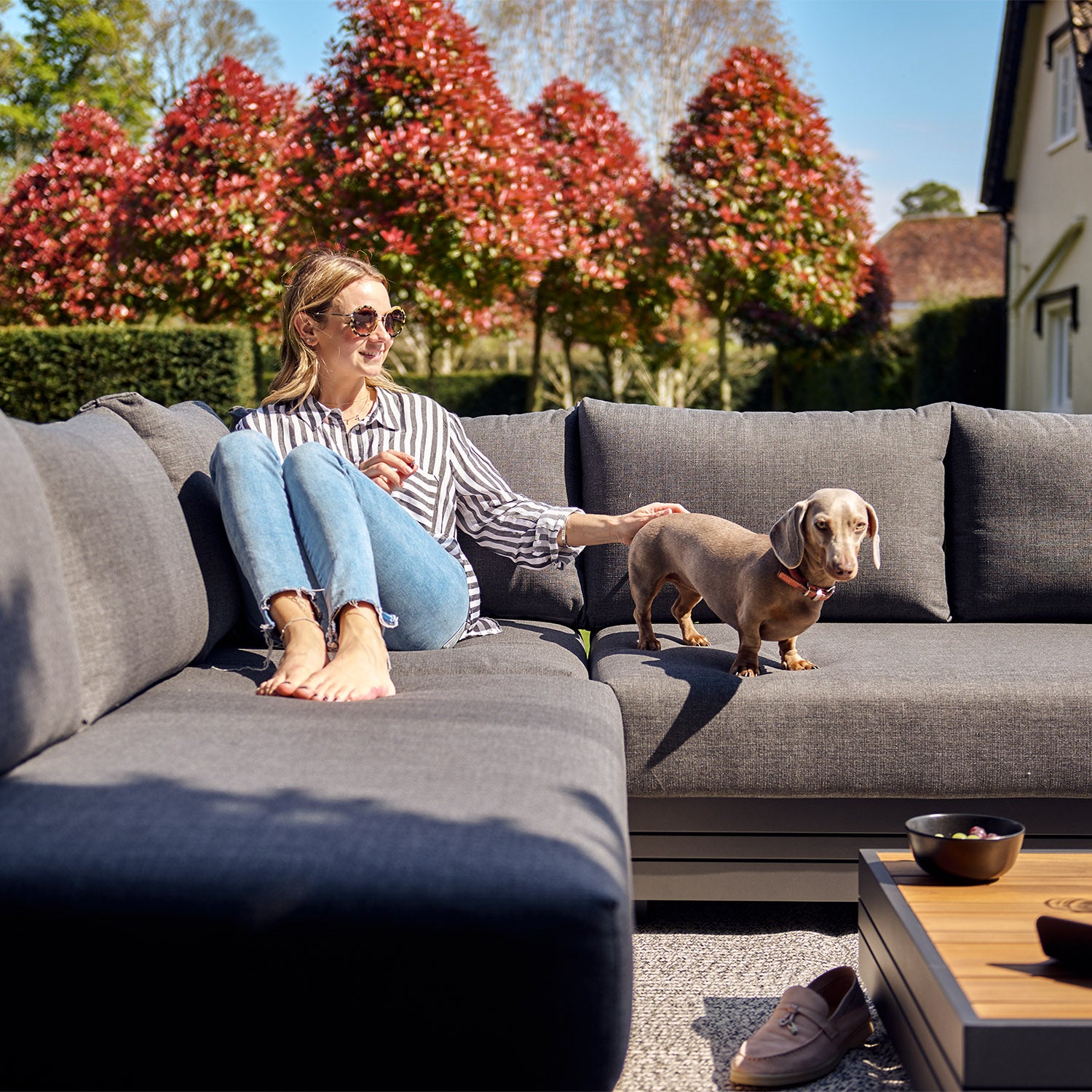 A woman in sunglasses sits on an outdoor sofa in a garden, petting a small dog, enjoying the stylish comfort of her Panama Luxury Outdoor Corner Group Set in Charcoal made from sustainable teak.