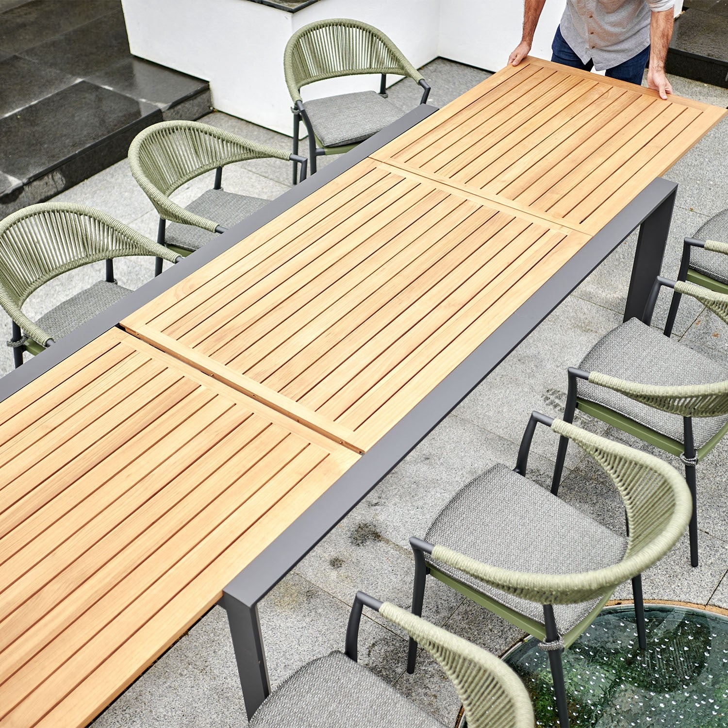 Person adjusting the Pier Extending Dining Table with an elegant teak top and aluminium leg frame, surrounded by green chairs with woven backs on a stone patio.