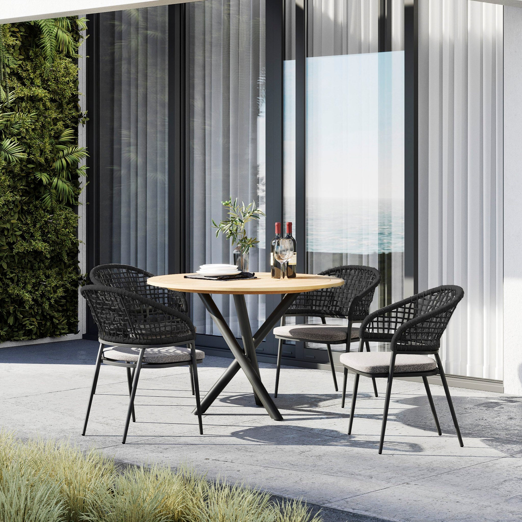 A modern outdoor patio set by large windows featuring the Kalama 4 Seat Round Dining Set with a Teak Table in Charcoal, four black woven chairs, and a bottle of wine with glasses.