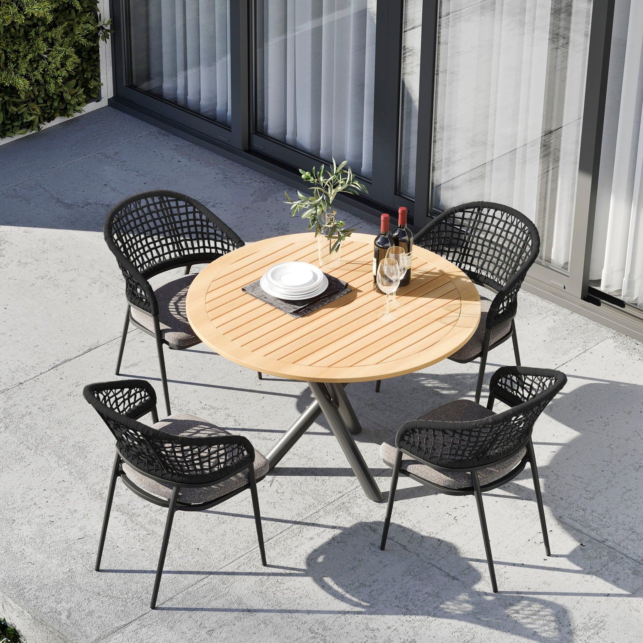 Kalama 4 Seat Round Dining Set with Teak Table in Charcoal featuring two wine bottles and a small plant, arranged on a concrete terrace.