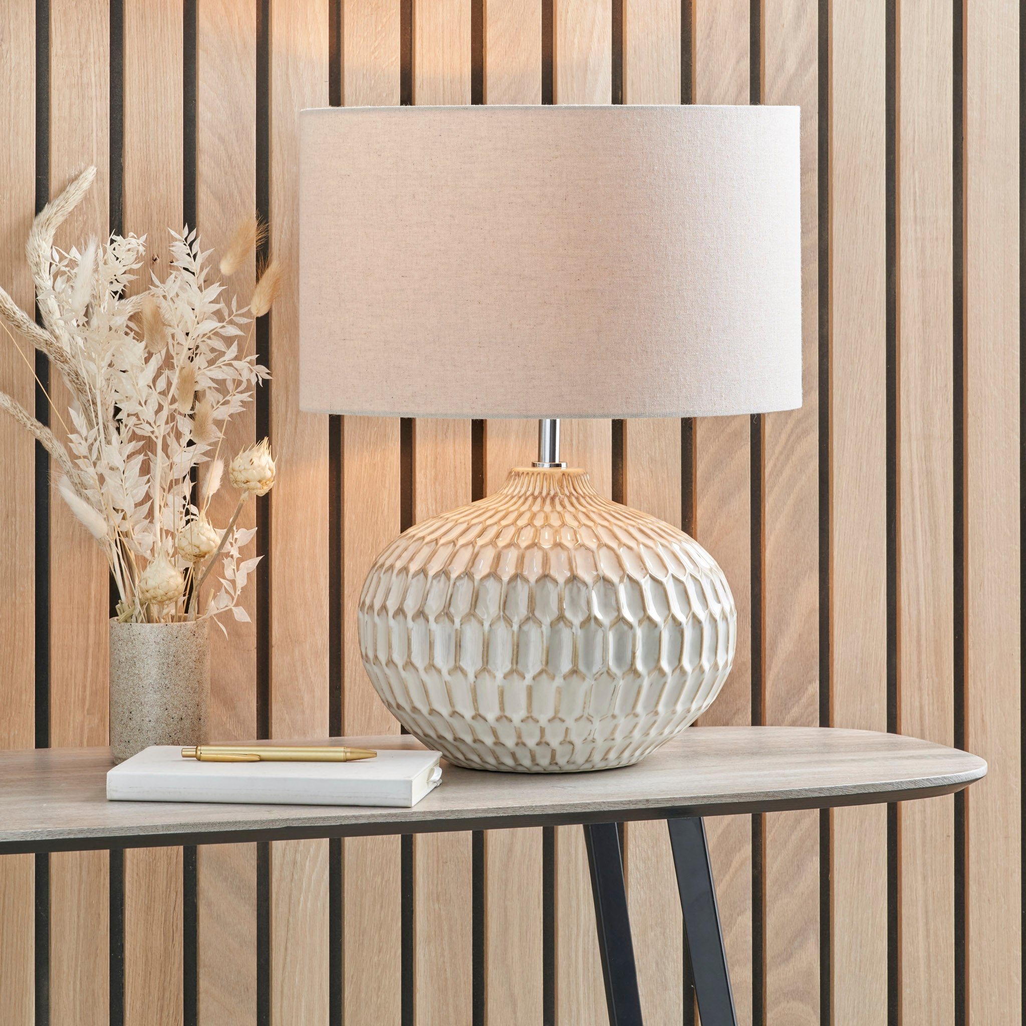 The Cassius Warm White Textured Glazed Ceramic Table Lamp with an Aztec texture and a linen lampshade is placed on a table decorated with dried flowers and a notebook.