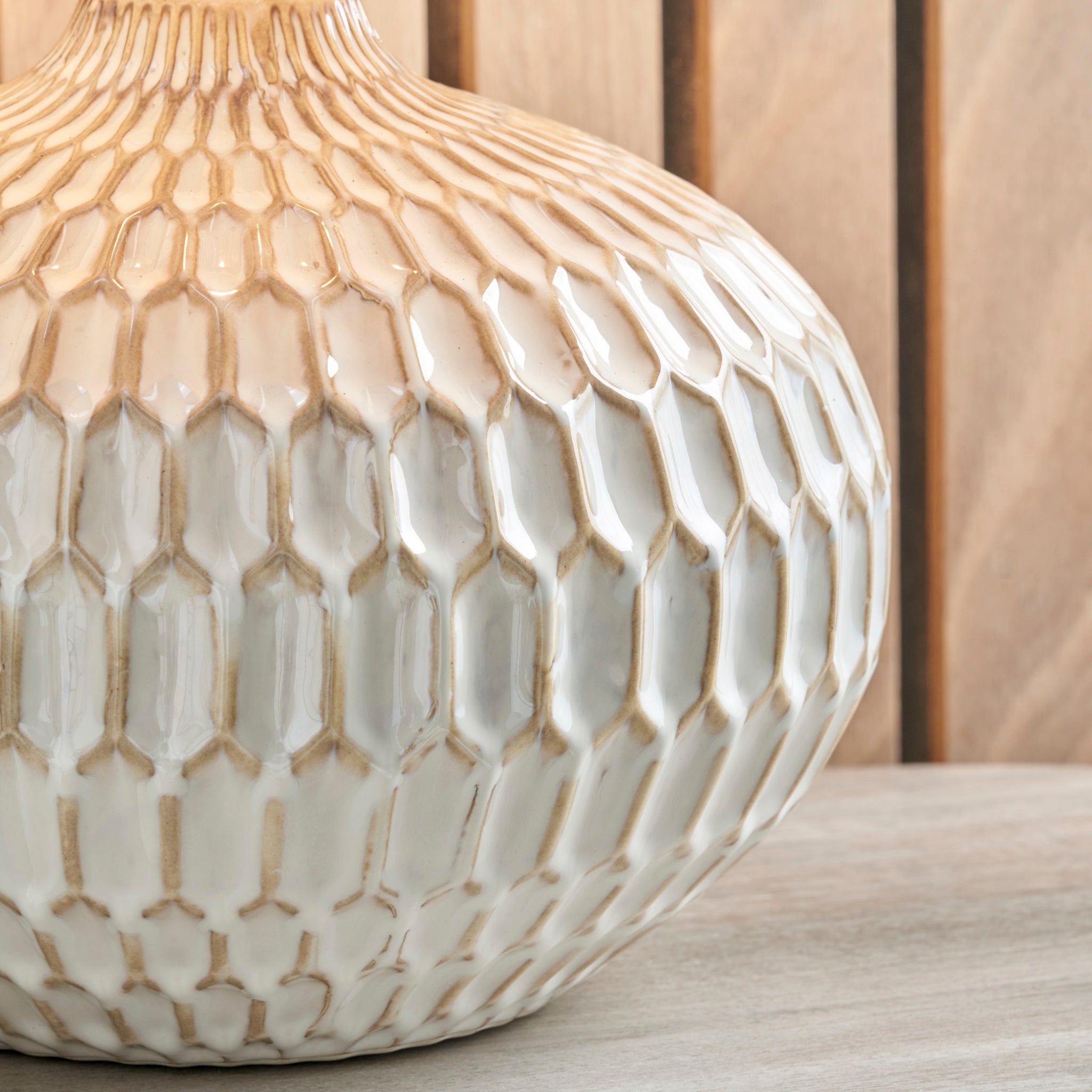 A close-up of the Cassius Warm White Textured Glazed Ceramic Table Lamp, showcasing its honeycomb pattern reminiscent of Aztec textures, is set on a wooden surface.