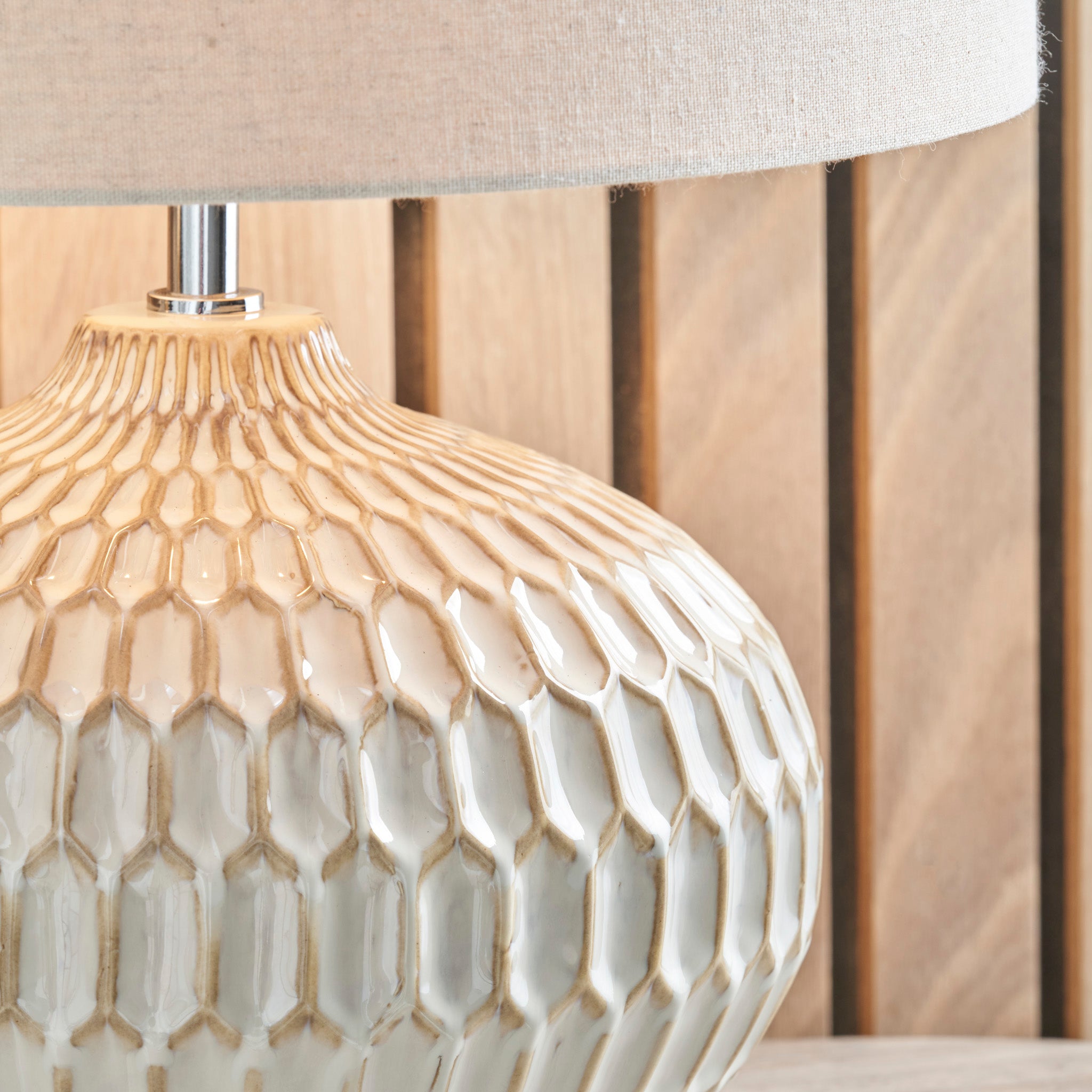 Close-up of the Cassius Warm White Textured Glazed Ceramic Table Lamp base featuring an Aztec pattern, with a beige linen lampshade, set against a wooden panel background.