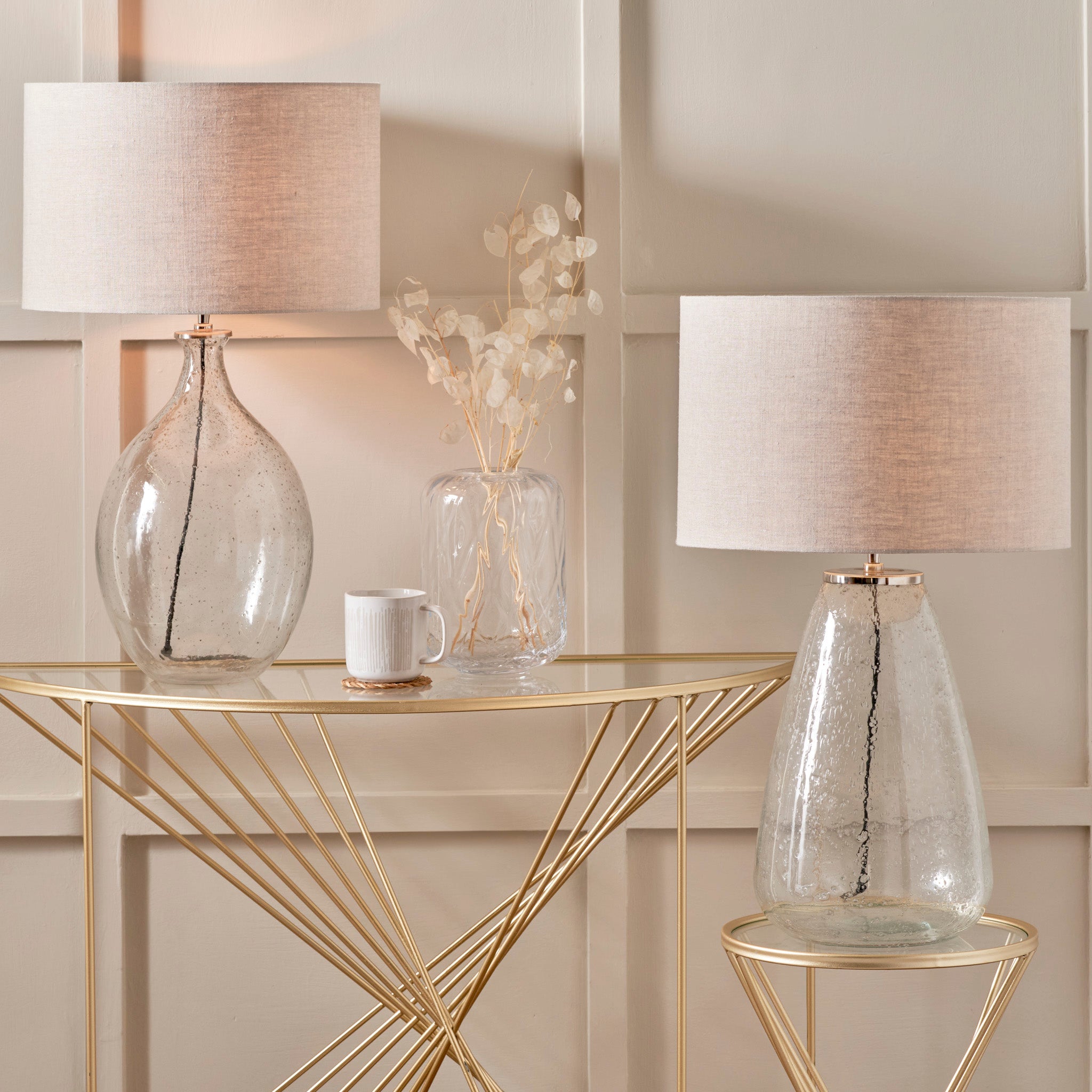 Two Elysia Bubble Glass and Silver Metal Curved Table Lamps with textured linen lampshades rest on gold tables, alongside a white mug and a clear vase with dried flowers.