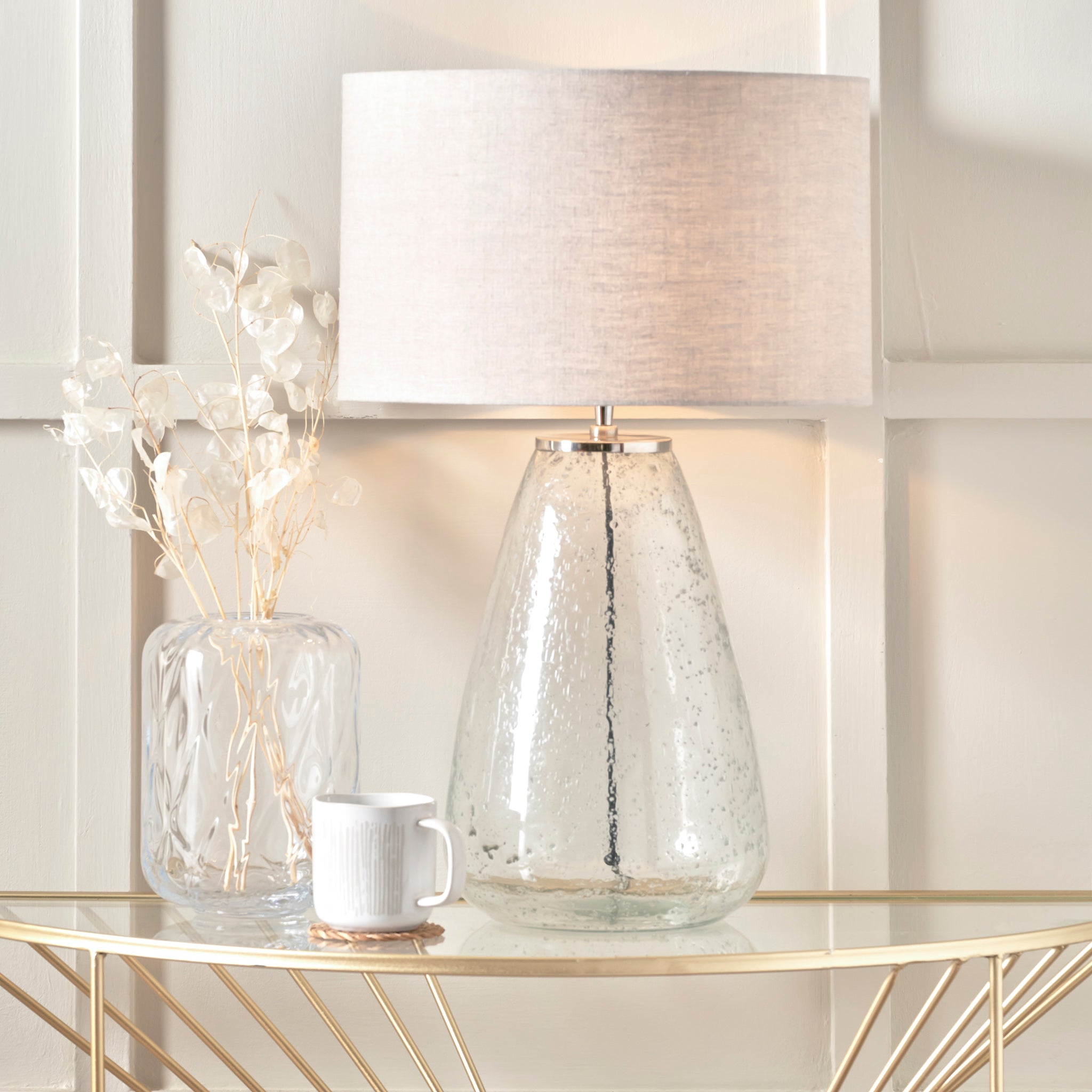 An Elysia Bubble Glass and Silver Metal Table Lamp with a natural linen lampshade stands elegantly next to a vase of dried flowers and a white mug on the decorative table.