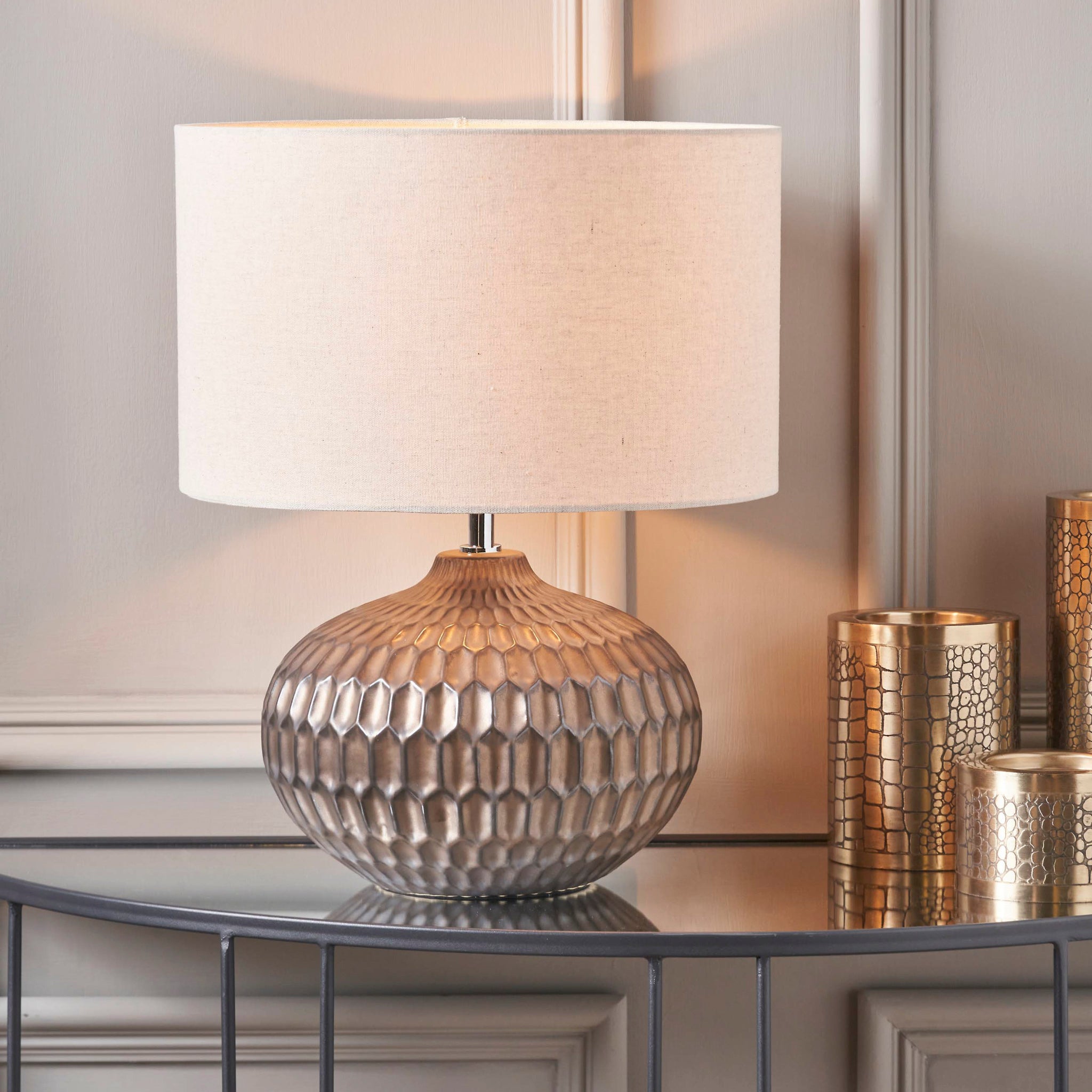 The Cassius Bronze Textured Glazed Ceramic Table Lamp with a linen-look shade sits beside gold candle holders on a glass console table.