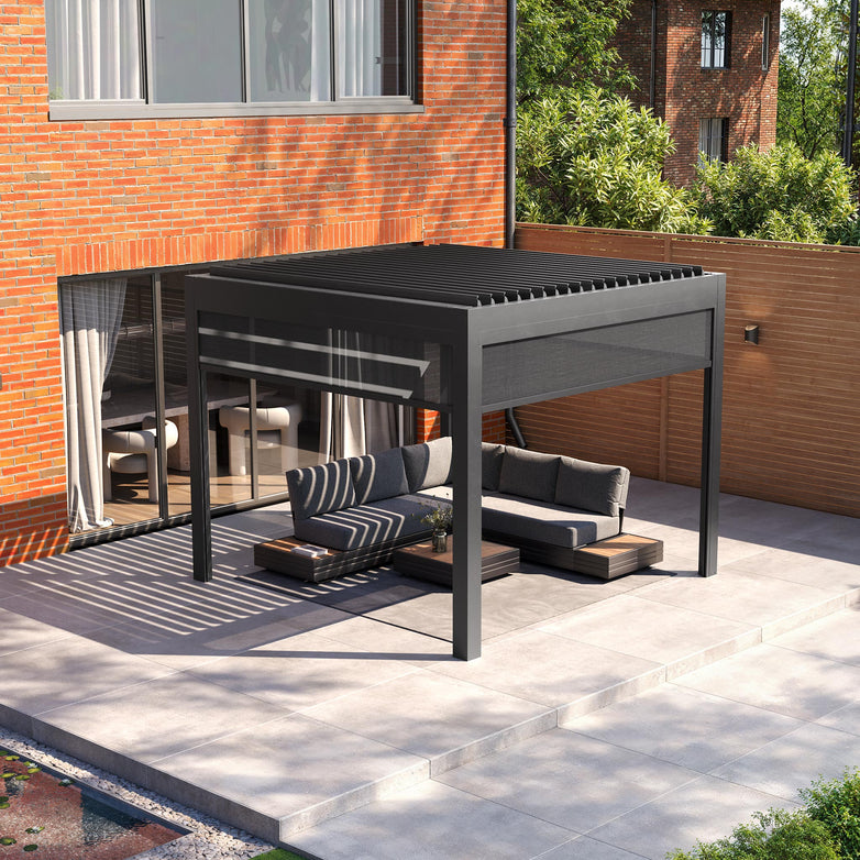 The PergoSTET Deluxe 3m x 3m square pergola with motorised LED louvres and three manual side blinds in grey complements a sunny patio featuring a sofa and table, situated next to a brick building with large windows.